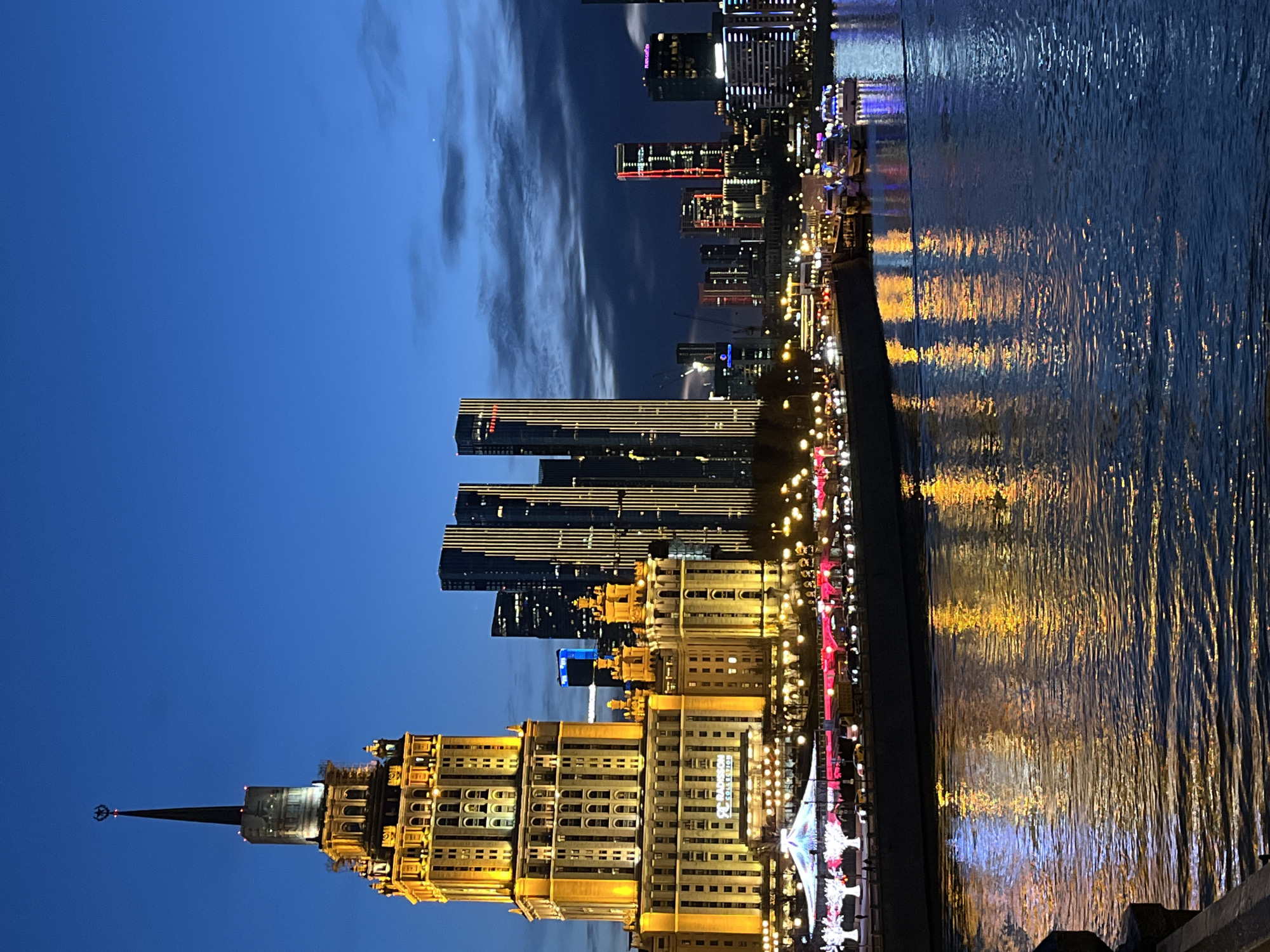 The image shows a nighttime view of Moscow, Russia. The Hotel Ukraina, one of the "Seven Sisters" skyscrapers, is prominently featured, illuminated in yellow light. Several modern high-rise buildings are visible behind it, also lit up at night. The cityscape is reflected in the Moskva River, with the reflections creating a shimmering effect. A row of lights lines the embankment along the river. The sky is dark blue with some wisps of clouds.