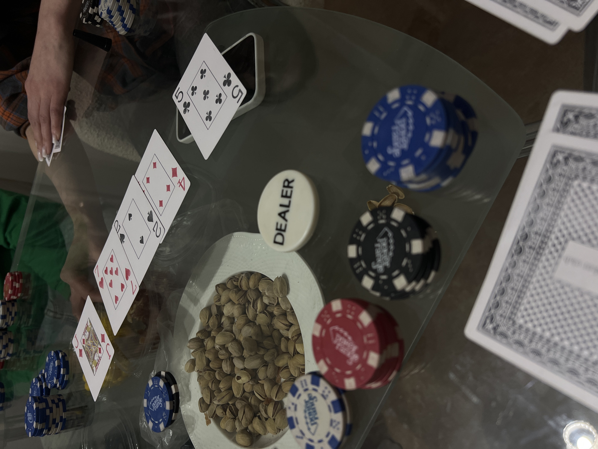 The image captures a casual indoor setting, likely a home, where a card game, possibly poker, is in progress on a glass table. A person's hand is visible on the left, holding a card, wearing a plaid shirt over a green garment.
On the table, several playing cards are laid out: a Five of Clubs standing upright, partially propped against a smartphone; a Four of Diamonds and a Queen of Spades (partially visible) laid flat; a Jack of Diamonds, a Seven of Hearts, and a Ten of Hearts (both partially visible) are also face-up. A stack of face-down cards, likely a deck, is to the right.
Various poker paraphernalia are scattered across the table. Stacks of poker chips in blue, red, and black are visible, some bearing the text "Poker Range." A white, circular "DEALER" button is placed near the center. A white bowl filled with pistachios sits prominently on the table, suggesting a snack during the game. The overall scene conveys a relaxed, social atmosphere. No specific time of day or outdoor weather is discernible, but the lighting suggests an evening or nighttime activity. The location context is Svkh Voronezhskiy, Russia.