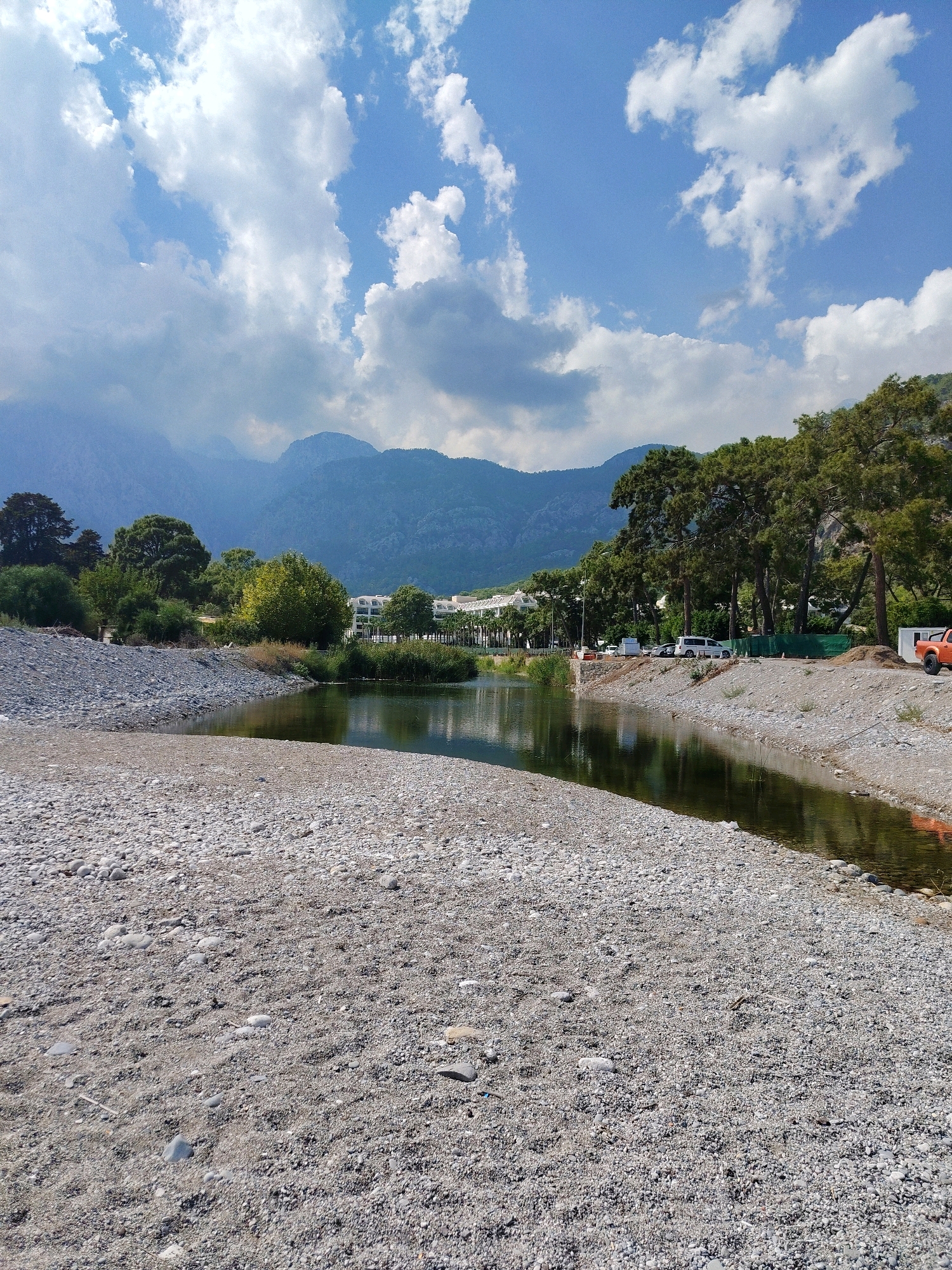 The image shows a tranquil scene by a river in Kemer, Turkey. The foreground is dominated by a wide, pebble-strewn beach that slopes gently down to the water's edge. The river itself is calm and reflects the sky and surrounding trees. On the opposite bank, lush green trees line the water, and beyond them, a modern white building with multiple balconies is visible, suggesting a resort or hotel. Further in the background, majestic mountains rise, shrouded in a soft haze, with a dramatic blue sky dotted with fluffy white clouds above. A few vehicles, including a white van and an orange utility vehicle, are parked near the tree line on the right bank, indicating human presence and development in the area. The overall atmosphere is peaceful and scenic, highlighting the natural beauty of Kemer.