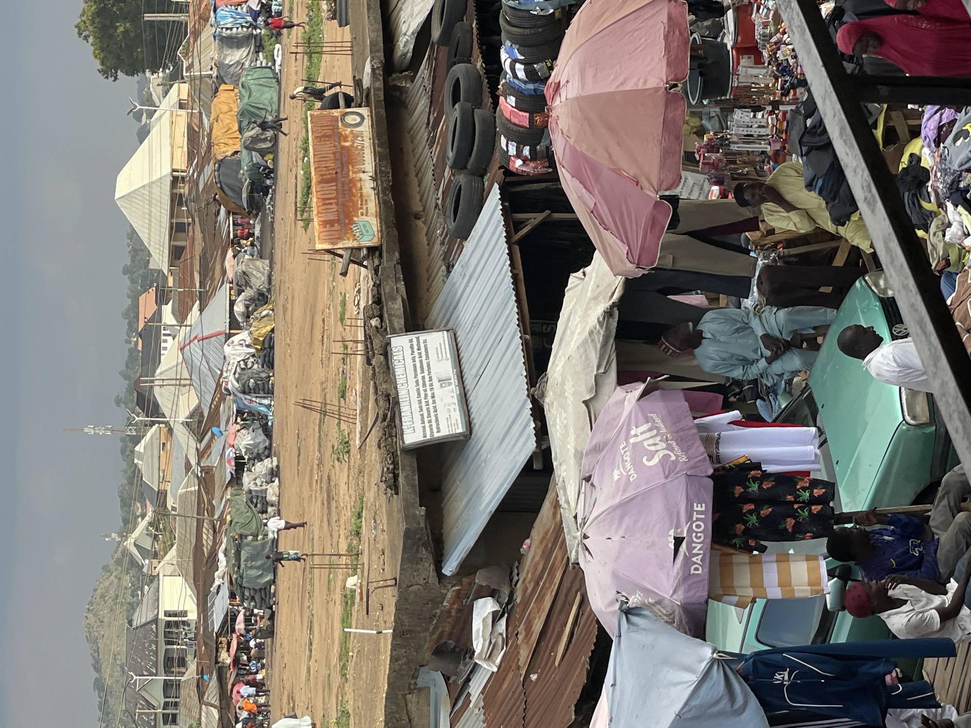 The image depicts a crowded marketplace in Bukuru, Nigeria. The scene is filled with a mix of residential and commercial buildings, many with corrugated metal roofs. A dusty, open area stretches between buildings, where people are visible engaging in commerce.
Several individuals are seen sitting or standing, some under umbrellas labeled "DANGOTE Salt Refined & Iodized," suggesting salt vendors. Some appear to be examining or selling merchandise displayed on vehicles, possibly car bonnets or other surfaces. A stack of tires is also visible.
Signage is evident, including one reading "DE MOTOR" and another with the label "K-FRANKLIN CHEMICALS," listing various chemicals. The overall impression is of a bustling, informal market environment with a mix of goods and activities. The weather appears overcast.