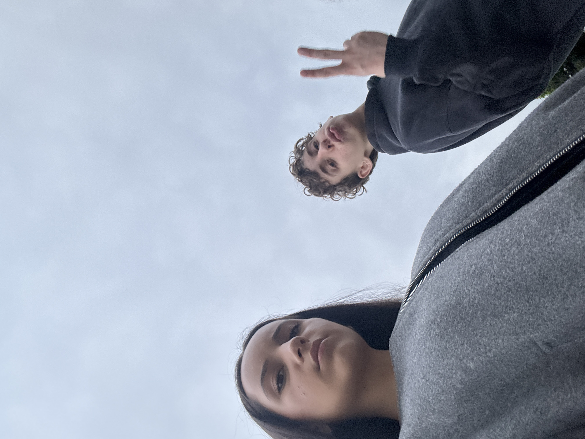 This low-angle outdoor photograph captures a young woman and a young man against an overcast sky. The woman, positioned on the left, has long, dark hair and is wearing a grey zipped jacket. She is looking directly at the camera with a neutral expression. To her right, the young man has curly brown hair and is wearing a black hooded sweatshirt. He is looking towards the camera with his lips puckered and is making a peace sign with his right hand. The background is dominated by a cloudy sky, with a hint of green foliage visible at the bottom right edge of the frame. The perspective suggests the photo was taken from a low vantage point, possibly looking up at the subjects.