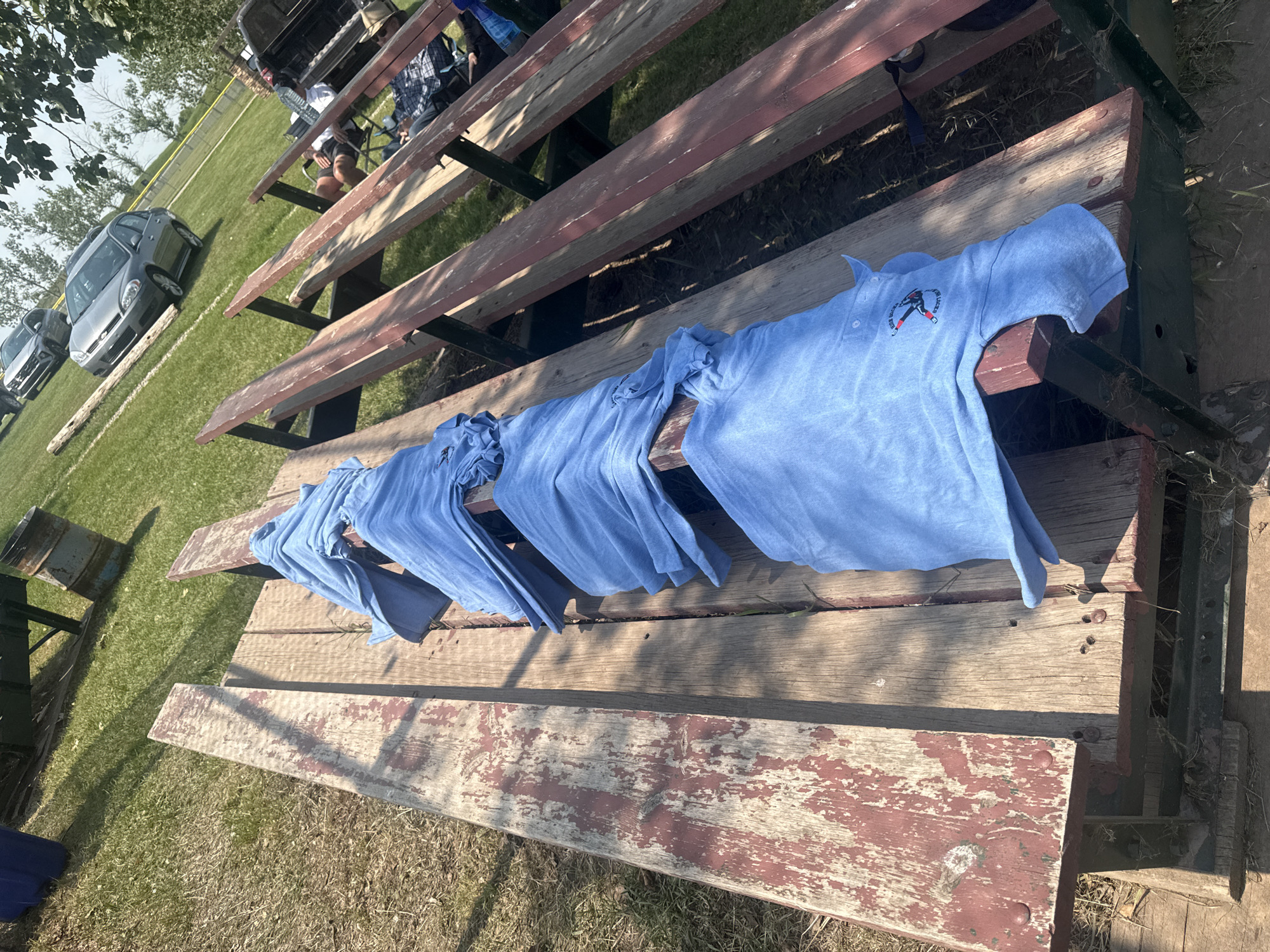 In AB, Canada, on a sunny day, blue shirts are laid out on a weathered wooden bleacher. The bleacher has a red and worn paint finish. In the background, green grass and trees are visible. Further back, several cars are parked on a paved area. Two individuals are seated on a lower bleacher, one wearing a hat.