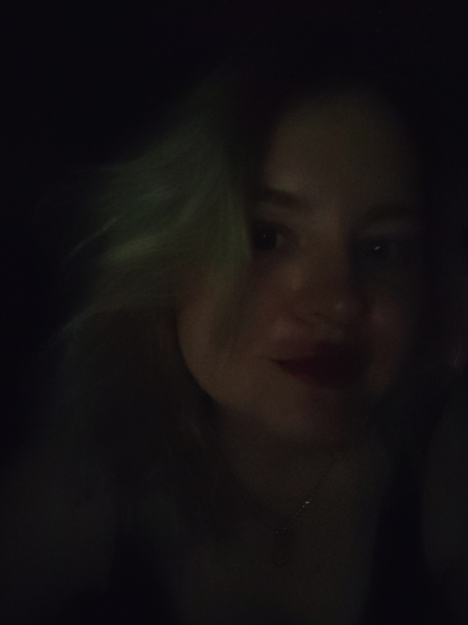 A dimly lit image shows the face of a woman with blonde hair. She appears to be wearing lipstick and a necklace. The background is dark and indistinct. The location cannot be determined from this image.