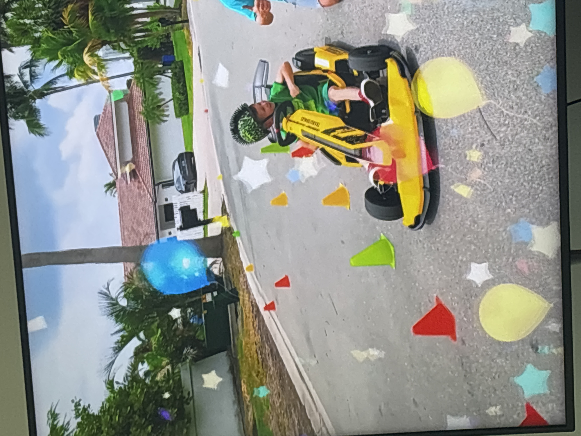 The image shows a young boy in a green shirt and a mohawk-style helmet driving a yellow go-kart. He is on a paved street lined with palm trees and houses. There are several colorful cones and balloons scattered on the street and in the air, some of which appear to be digital overlays. In the background, a dark car is parked in front of a house with a tiled roof. The sky is partly cloudy. The scene depicts a playful or celebratory event. This image is not located in Dubai, United Arab Emirates.