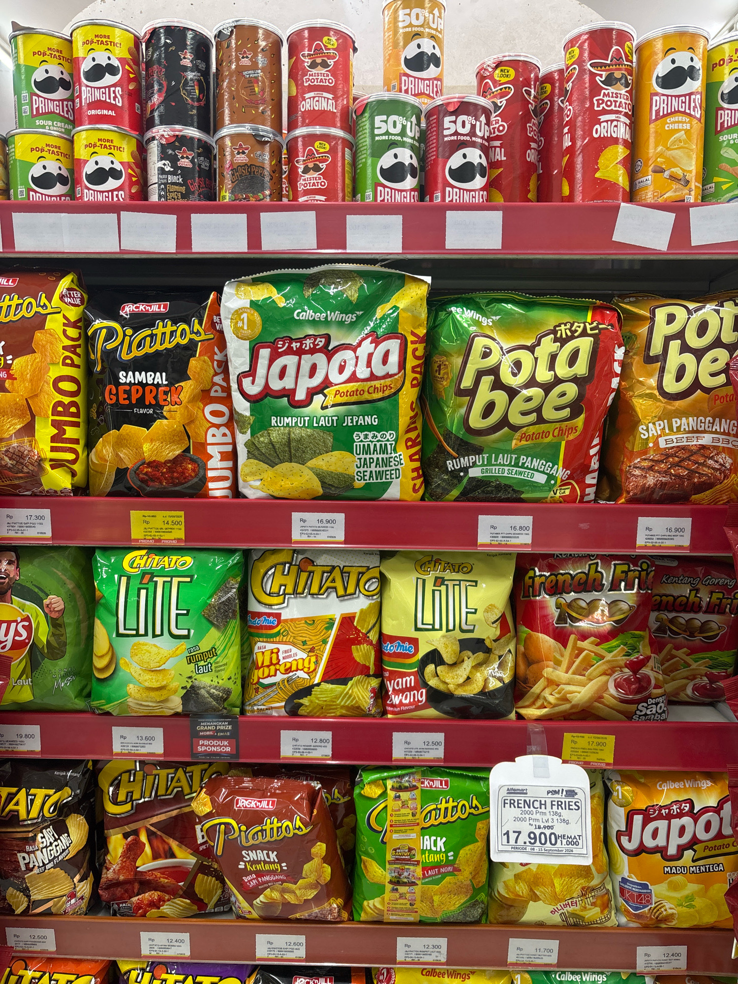 This is a retail product photograph taken in a supermarket, specifically at Alfamart in Cilegon, Indonesia. The image displays multiple shelves stocked with various snack products, predominantly potato chips and similar items. The products are arranged in an organized manner, with labels and pricing clearly visible for many of them.
On the top shelf, there are several cans of Pringles chips in different flavors like Original, Sour Cream & Onion, and Cheese. Alongside the Pringles are cans of "Mister Potato" chips, also in various flavors.
The middle shelf features an assortment of bagged snacks. These include "Jumbo Pack" chips, "Piattos" chips with "Sambal Geprek" flavor, and "Potabee" potato chips in "Grilled Seaweed" and "Beef BBQ" flavors. There are also bags of "Chitato Lite" chips with "Seaweed Flavor" and "Fried Noodles" flavored snacks.
The bottom shelf continues with more bagged snacks. We see "Chitato" chips in "Beef BBQ flavor" and a spicy flavor with chicken. "Piattos" in "Roasted Chili" flavor is also present. Further down, "French Fries" snacks and more "Japota" potato chips are visible. The overall environment suggests a well-lit retail space focused on packaged food items. The pricing labels are mostly in Indonesian Rupiah (Rp).