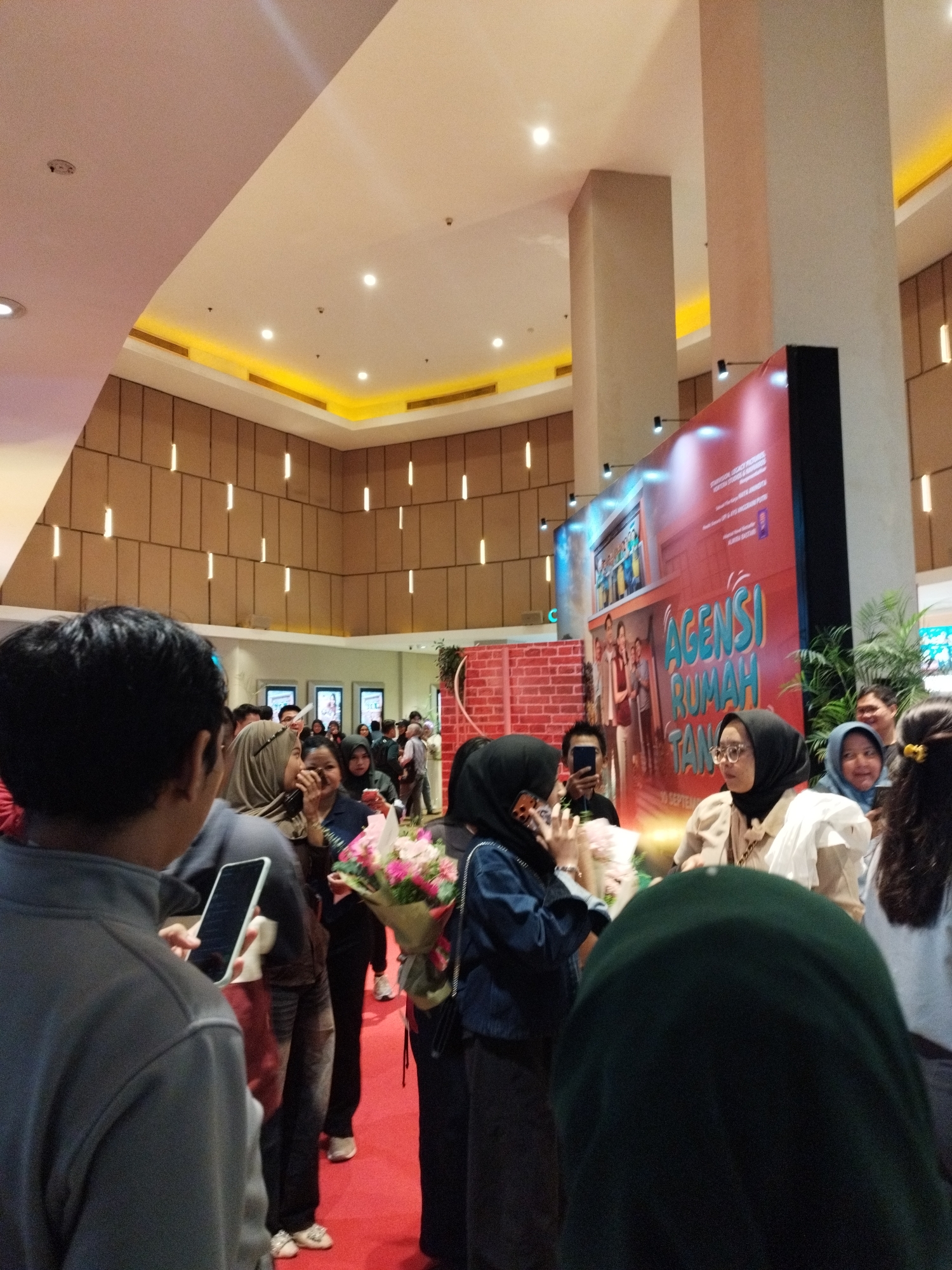 The image depicts an indoor gathering, likely at a cinema or event venue in South Jakarta, Indonesia, as indicated by the presence of a large screen displaying "AGENSI RUMAH TANGGA" and a red carpet. A group of people, predominantly young adults and some children, are present, many wearing headscarves. Some individuals are holding smartphones, presumably taking pictures or videos. A woman in the center is holding a bouquet of flowers, suggesting a celebratory or welcoming occasion. The background features architectural elements like columns and a patterned wall with vertical lighting. The lighting appears to be a mix of ambient and spotlighting, common in such venues.