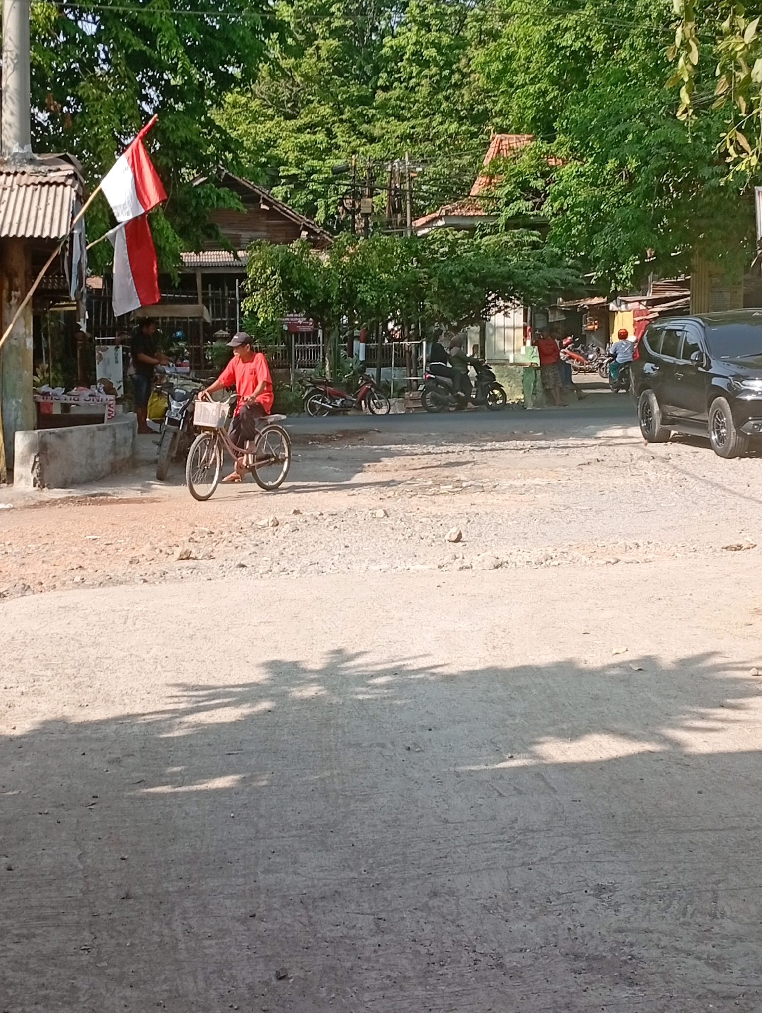 The image depicts a street scene in Jogosari, Indonesia, bathed in sunlight. In the foreground, a dusty, unpaved road shows scattered pebbles and debris, with tree shadows cast across its surface. On the left side of the road, a structure with a corrugated metal roof is visible, adorned with two Indonesian flags on poles. A person in a red shirt is seated on a bicycle with a basket, positioned on the unpaved area of the road. Behind the cyclist, a motorcycle is parked. In the background, several people are gathered near what appears to be a small shop or stall. Beyond them, there are more motorcycles and a dark-colored SUV parked on the paved portion of the road. The background is filled with lush green trees and buildings with tiled roofs, suggesting a residential or mixed-use area.