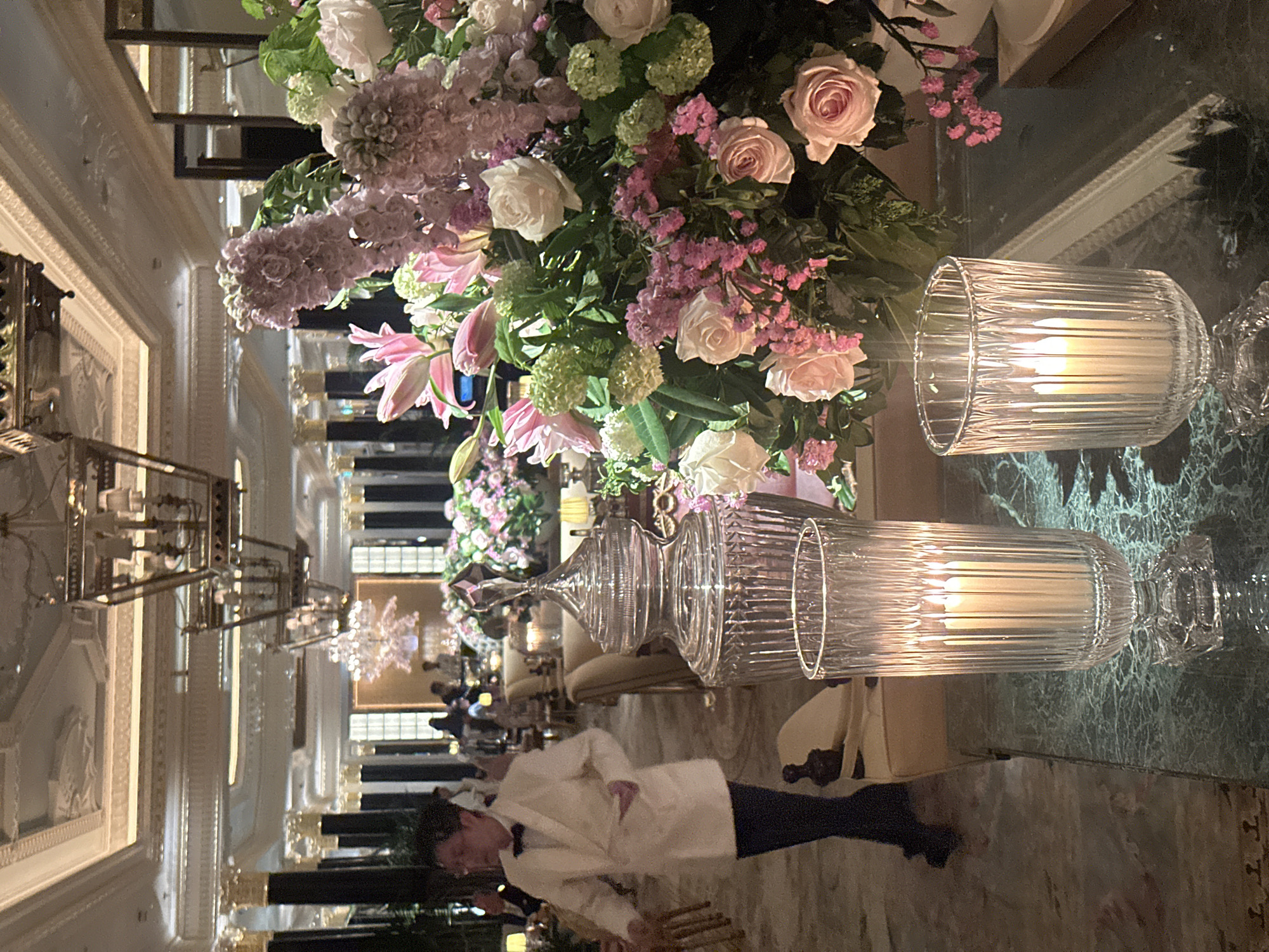 The image captures an interior setting, likely within a luxurious hotel or event space in London, United Kingdom. A table arrangement is prominent, featuring a lush floral centerpiece composed of pink and white roses, hydrangeas, lilies, and other flowers. Two large, ornate glass candle holders with lit candles are placed near the floral arrangement.
In the background, there is a view through the space revealing additional tables, chandeliers, and other patrons. In the foreground, there is a person wearing a white jacket and black trousers standing.