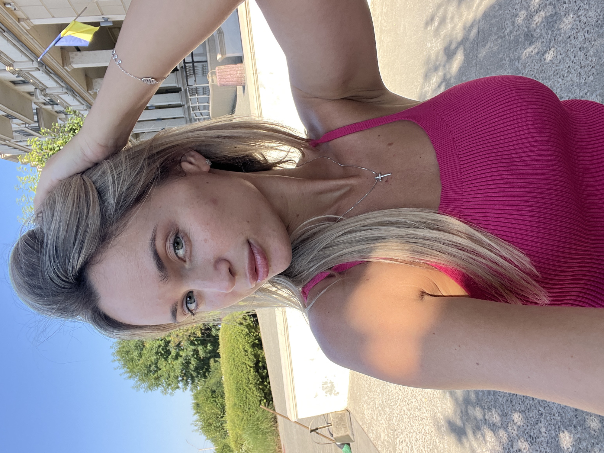 A blonde woman with fair skin, her hand in her hair, poses for a selfie outdoors on a sunny day. She is wearing a bright pink ribbed tank top and a silver necklace with a cross pendant. A bracelet with several charms adorns her left wrist. In the background, a building with a Ukrainian flag flying from its facade is visible, suggesting the location is in Ukraine. Green foliage and a blue sky complete the scene.