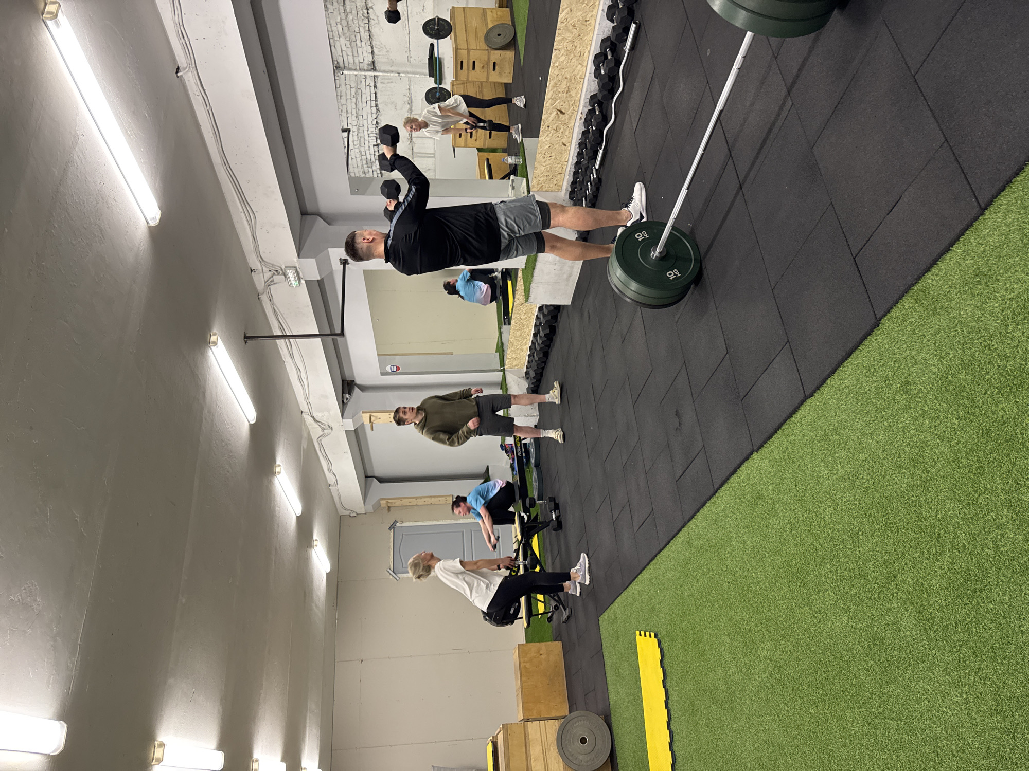 This image depicts an active indoor fitness gym or CrossFit facility located in Chaikovsky, Russia. The space is well-lit by numerous linear fluorescent tube lights mounted on the light grey walls and ceiling, some of which feature exposed brick textures.
The gym floor is distinctly divided between black rubber interlocking mats and a vibrant green artificial turf area, separated by a diagonal line. Several individuals are engaged in various workout activities. In the foreground on the black mats, a man is seen from behind, wearing a black long-sleeve top and grey shorts. He holds two black hexagonal dumbbells raised above his shoulders, indicative of a pressing exercise. Beside him, a barbell rests on the floor, loaded with two green plates, each marked "10 kg."
Further back on the black mats, a woman in a white t-shirt and black leggings appears to be bending over or lifting an object near a wooden plyometric box. Nearby, another man in an olive green hoodie and dark shorts stands on the edge of the artificial turf, looking towards the left. In the background, a person in a blue t-shirt is actively using a rowing machine, while another woman with light hair is seated on a piece of equipment, possibly resting or preparing for an exercise.
The gym is equipped with various fitness gear, including racks of dumbbells along a wall, additional wooden boxes, and bright yellow interlocking mats on the green turf. The scene suggests a focused and energetic workout session.