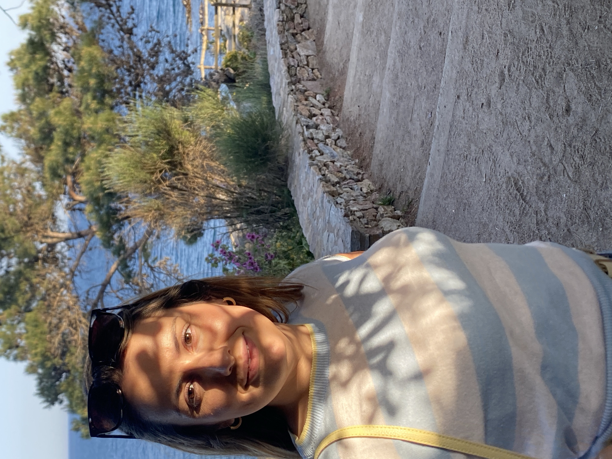 The image depicts a woman in a striped top, wearing sunglasses, standing on stone steps with a stone wall to the right. In the background, there is a view of the sea, trees, and vegetation. The location is Calella De Palafrugell, Spain, given the typical scenery and architectural elements present.