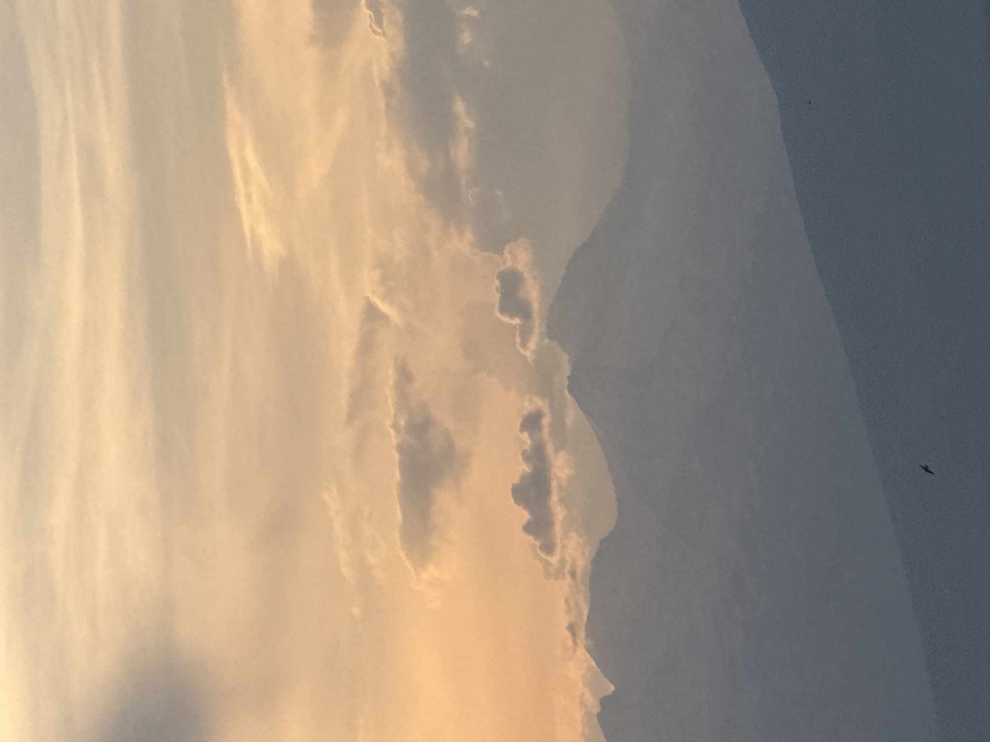 This is a vertical, medium-shot photograph of a mountainous landscape at sunset. The sky is a warm gradient of peach and pale yellow, with streaks of light orange and hints of gray clouds scattered throughout. The clouds are mostly thin and wispy, with a few darker, more defined formations visible. In the middle ground, a series of silhouetted mountain peaks rise against the colorful sky. These mountains are rendered in shades of gray and blue, with subtle undulations suggesting their rugged terrain. The foreground is dominated by a dark blue, sloping hill, which is partially obscured by shadow. A small, dark bird is captured in flight in the lower left quadrant of the image, positioned over the sloping hill. The overall impression is one of serene natural beauty at the end of the day. The location is Abovyan, Armenia.