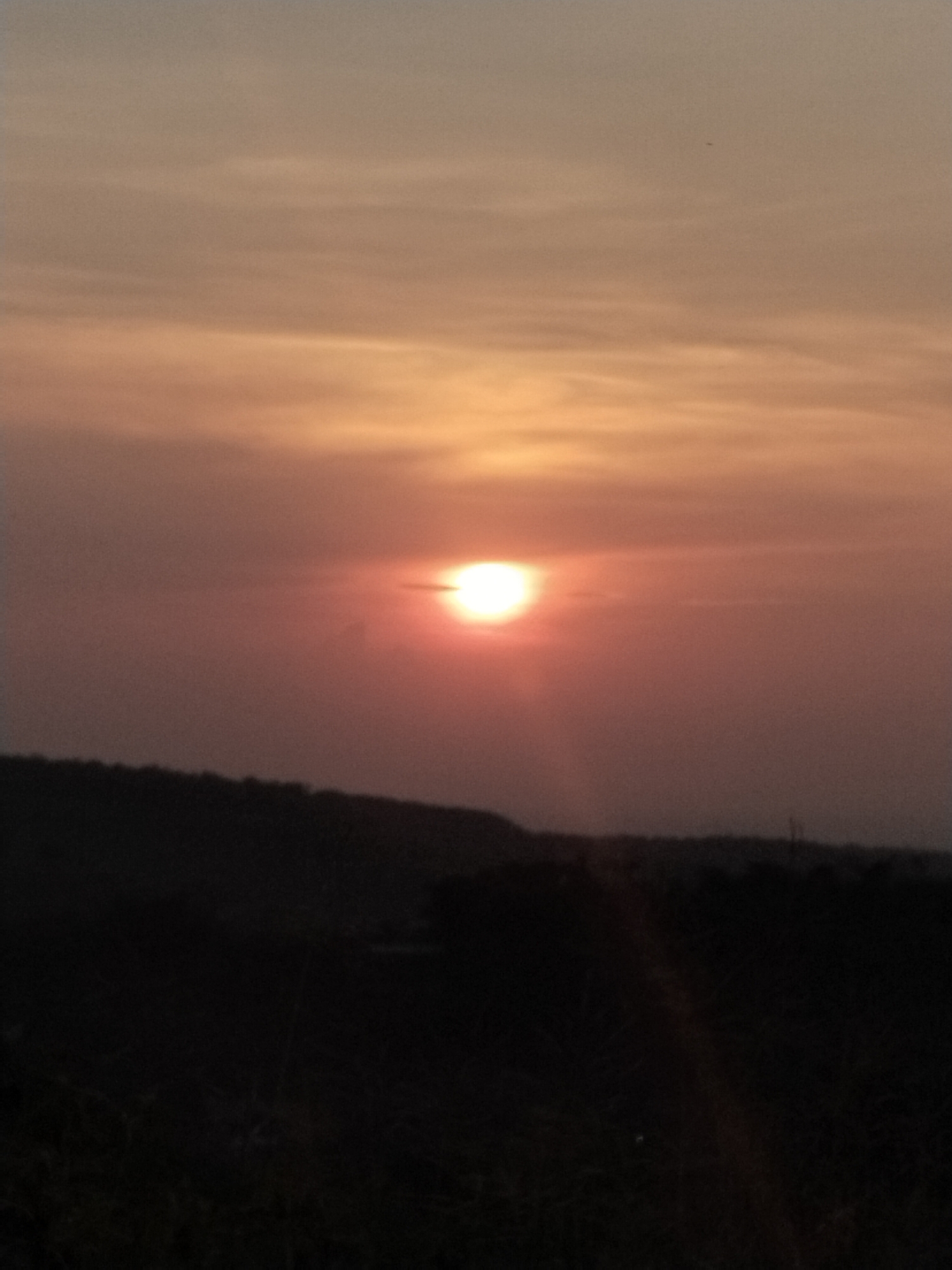 The image displays a serene sunset scene over a natural landscape. The primary subject is the sun, appearing as a bright, glowing red-orange disc centered in the upper mid-frame. Its light is diffused by a hazy or cloudy sky, creating a soft, warm glow that transitions from vibrant orange and yellow closer to the sun and horizon, gradually fading into muted browns and grays higher in the sky. Subtle, elongated cloud patterns are visible across the sky.
Below the horizon, a dark, undulating silhouette of distant hills or mountains stretches across the frame. The foreground is cast in deep shadow, presenting as an indistinct, almost black mass of vegetation or land, with minimal discernible details. A small, dark speck, possibly a bird or insect, can be observed against the lighter sky in the upper portion of the image.
The scene captures the twilight hour, with the sun low in the sky, indicative of either sunset or sunrise, though the overall dimness and color suggest sunset. The weather appears hazy or slightly overcast. The setting is an undeveloped, natural environment. While no specific geographical markers are visible within the image, the provided context places the location in Jambeyan, Indonesia. No visible text is present in the media. The image conveys a sense of peaceful, natural beauty.