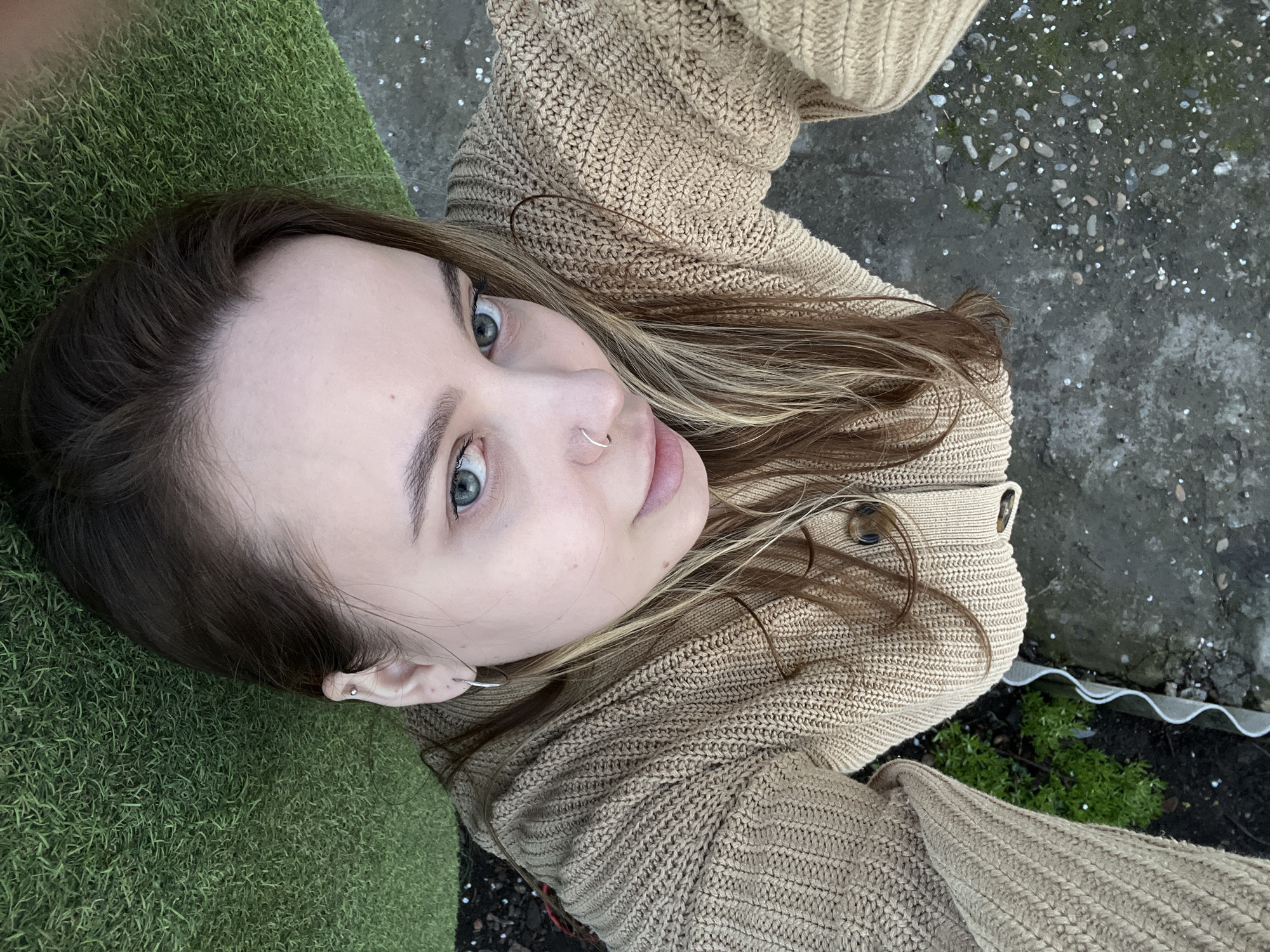 A woman with light skin and blue eyes is looking upwards at the camera. She has light brown hair with blonde highlights and a nose ring in her left nostril. She is wearing a tan knit sweater. The background consists of green artificial turf, a concrete surface with small pebbles, and vegetation. The location is not specified as Novopokrovskaya, Russia, and is not discernible from the image.