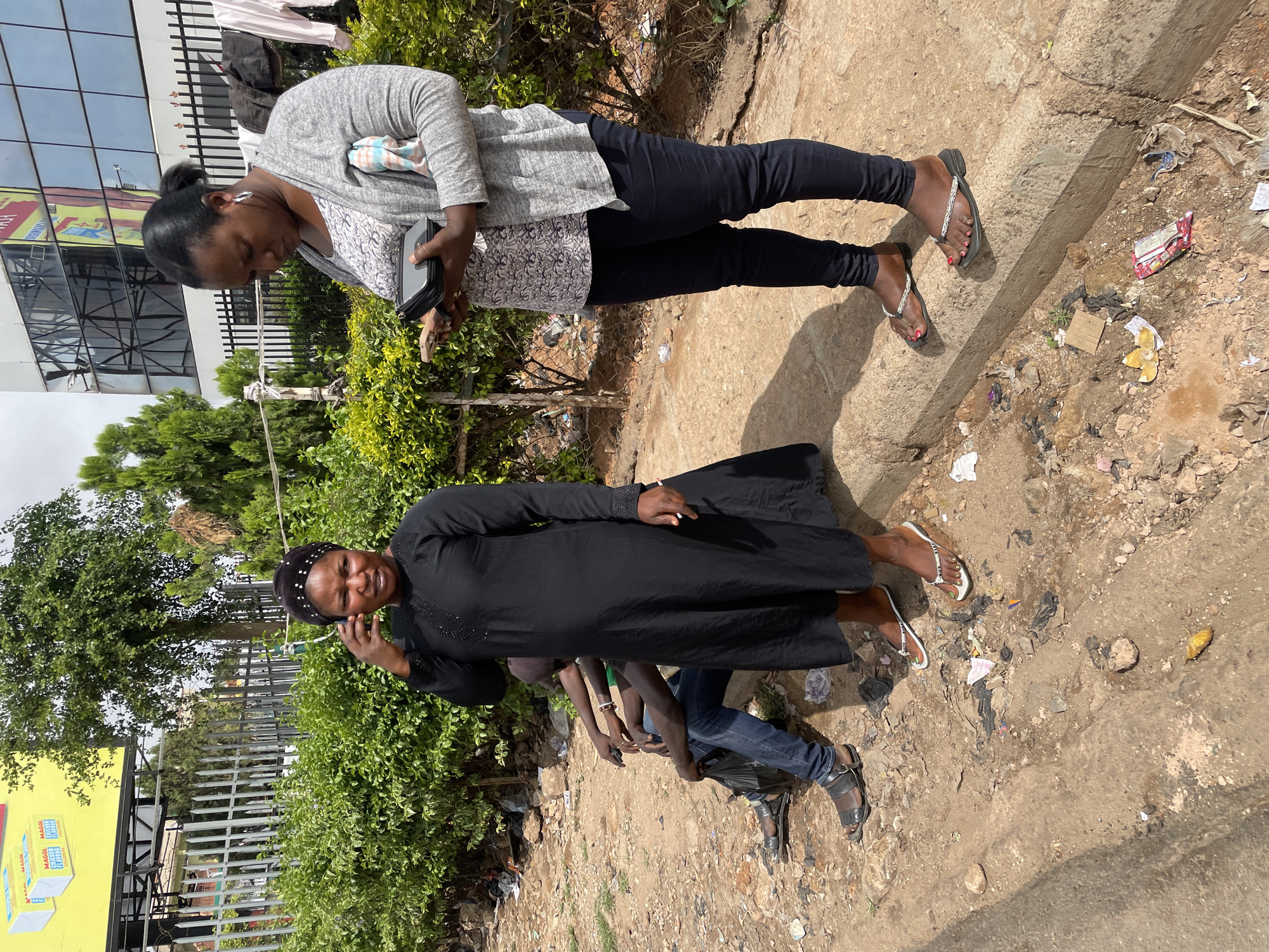 Two women stand on a cracked concrete sidewalk bordered by a dirt and gravel area. One woman wears a gray sweater, patterned shirt, dark pants, and sandals and is looking at a black wallet or phone in her hands. The second woman is wearing a long black dress and a beaded head wrap. She is holding a cell phone up to her right ear, speaking on the phone. A third person, a man wearing jeans and sandals is partially visible behind the second woman. The scene has litter on the ground. A building with reflective windows, green trees, and a metal fence are also visible. A yellow billboard is mounted on the metal fence. Based on these details, the location is Jos, Nigeria.
