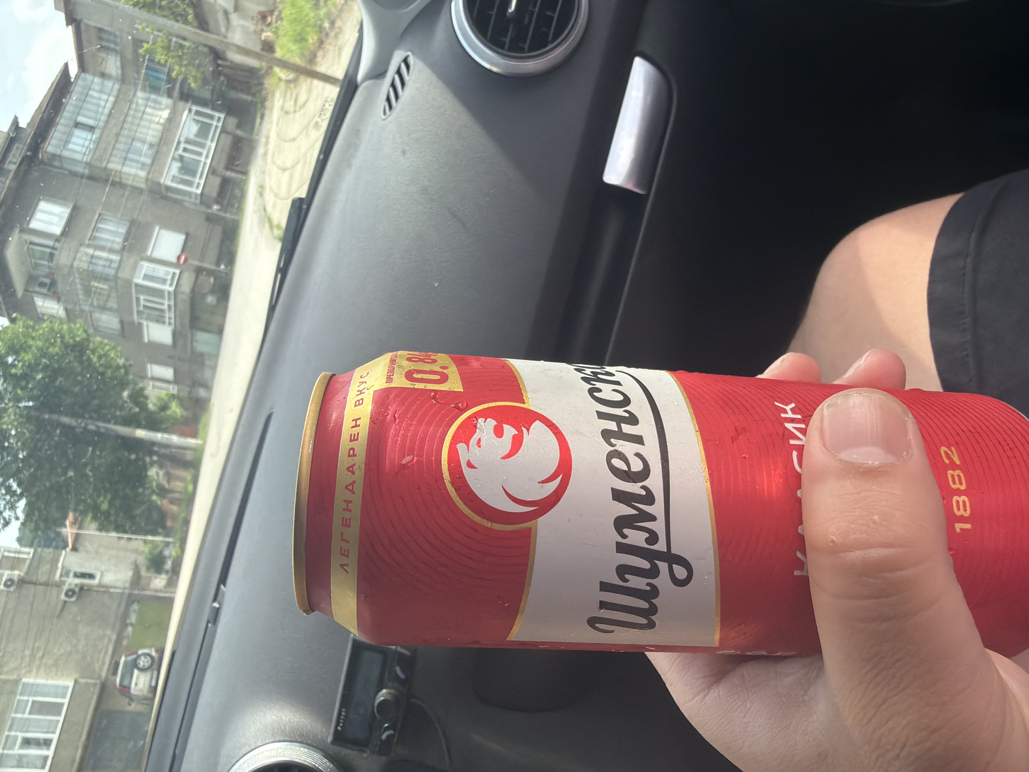 A first-person perspective photograph captures a hand holding a red and white can of "Шуменско" (Shumensko), a Bulgarian beer brand. The can displays "0.0%" indicating it's non-alcoholic, "ЛЕГЕНДАРЕН ВКУС" (Legendary Taste), "КЛАСИК" (Classic), and the year "1882," alongside a white winged creature logo on a red circle. Visible condensation suggests the can is cold. The hand has short fingernails, and the person appears to be wearing black shorts.
The setting is inside a car, with a black dashboard featuring air vents and a partially visible "Parrot" device. Through the car's windshield, an outdoor scene is visible on a bright, sunny day. The background features an older, grey apartment building with multiple windows and enclosed balconies, typical of Eastern European architecture. Green trees are seen along what appears to be a street or sidewalk, and a small portion of a white car is visible near the building. A faint red circular sign can be discerned on the building. The Cyrillic text on the can and the architectural style strongly suggest a location in Bulgaria, consistent with Shumen. The activity involves holding a cold drink, possibly while traveling or parked.