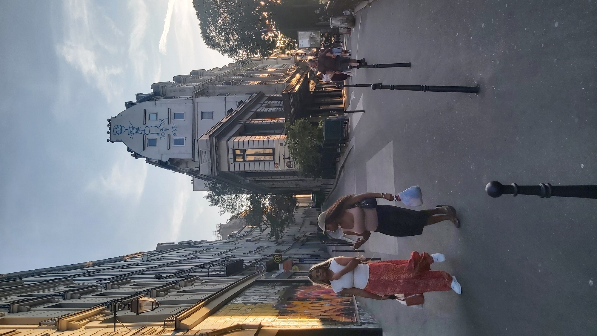 Two women, one in a white top and patterned skirt, the other in a tan top and dark skirt, walk down a cobblestone street in Paris, France. Both women are carrying shopping bags. The street is lined with buildings, one of which features graffiti. In the background, other pedestrians are visible, and the sun is setting, casting long shadows. The scene appears to be in a residential or commercial area of the city, possibly Montmartre given the architecture and artistic elements.
