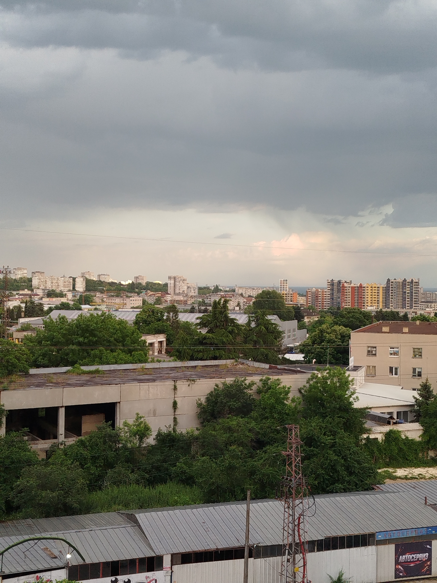 The image shows an elevated view of a cityscape, likely taken from a high vantage point in Varna, Bulgaria, on an overcast day with dark storm clouds filling the sky.
In the foreground, there is an industrial area. There are buildings with aging, flat roofs heavily overgrown with greenery, suggesting disuse or neglect. Below these structures, metal-roofed warehouses are visible. An electrical transmission tower is situated among the vegetation, with its red and silver structure adding an element of decay.
Beyond the foreground, the urban sprawl of Varna becomes apparent. Residential buildings of varying heights and colors dot the landscape, interspersed with green spaces, probably parks or gardens. The architectural style seems to be a mix of older, low-rise structures and newer, taller apartment blocks. In the midground, there are buildings with facades painted in multiple colors.
The background features a distant skyline, with indistinct forms suggesting a continuation of the city toward the horizon, where the sky is slightly brighter, possibly indicating the location of the Black Sea. The lighting is diffused due to the heavy cloud cover, casting a muted tone across the entire scene. There are no people present in the picture. A building with the Russian word "АВТОСЕРВИЗ" is visible.