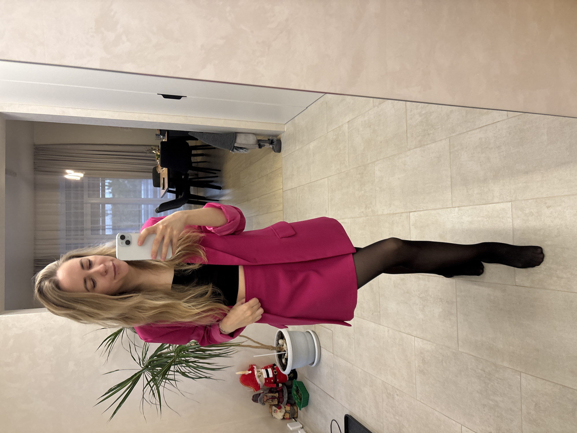A woman is taking a selfie in what appears to be an apartment. The woman has long blonde hair and is wearing a pink blazer, a black top, and black pantyhose. She is holding a white smartphone.
To the left of the woman, there is a potted plant with long, thin leaves. Underneath the plant, there are small decorative figurines.
In the background, a door is visible, leading into another room with a dining table and chairs. The floor appears to be tiled.
There is no evidence of the image location being in the city of Chaikovsky, Russia.