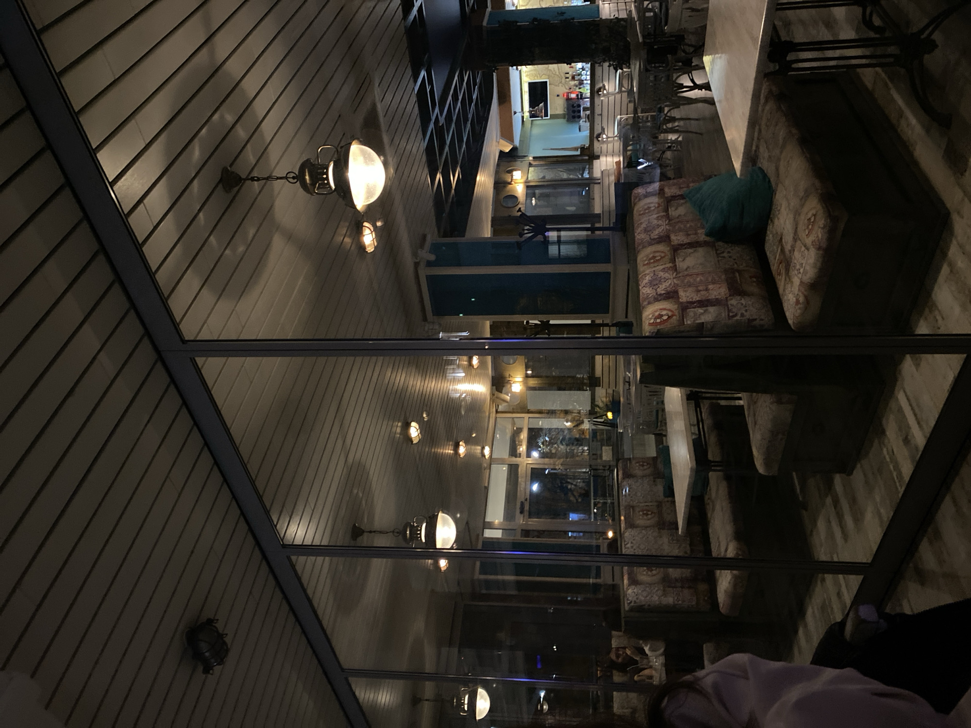 The image appears to be an interior shot of a restaurant or café. The location is suggested to be Stara Zagora, Bulgaria. The scene shows tables and cushioned seating areas with a decorative fabric pattern. Tables are set up, possibly for dining. The restaurant has a visible outdoor section or view, indicated by the windows showing external lights. There are various light fixtures on the walls and ceiling, casting warm light on the interior. There are people present, but the image does not provide clear faces or actions. The environment has a reflective surface, giving a mirrored view.