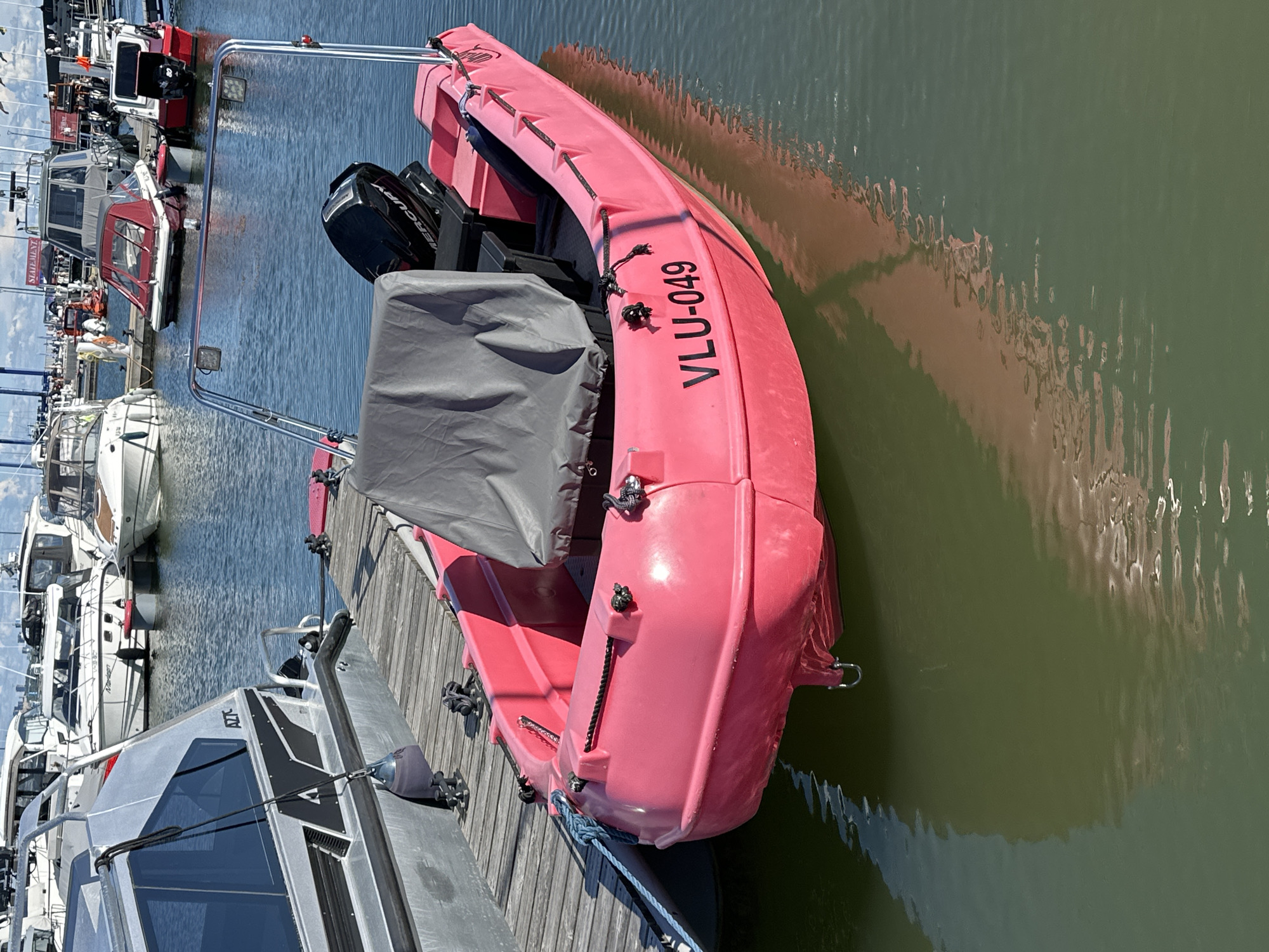 The image depicts a pink inflatable boat with the registration "VLU-049" docked at a marina. The boat is covered with a gray tarp, and a black Mercury outboard motor is visible at the stern. Other boats and the shoreline are visible in the background, suggesting a busy harbor environment. The water appears calm, reflecting the boats and the sky. The scene is likely located in Tallinn, Estonia, given the prevalence of this type of boat and marina infrastructure in the region.