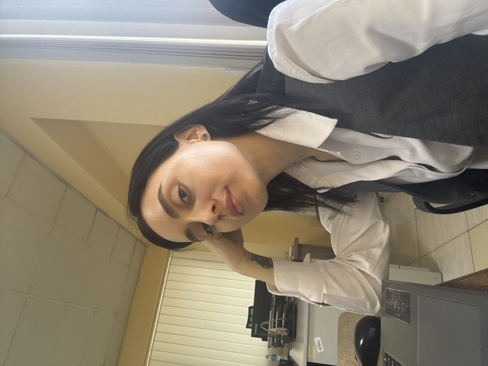A young woman with dark hair is posing for a photograph indoors. She is wearing a white collared shirt with the sleeves rolled up, and a dark vest over it. She has an earring in her left ear and a tattoo on her left arm. Her head is tilted slightly to the left, and she is sticking out her tongue.
She is seated at a desk. A Lenovo computer monitor is visible in the background. An office environment with beige-colored walls, tiled flooring, and horizontal blinds on the windows can be seen.
The image was likely taken in Tbilisi, Georgia, given the request.