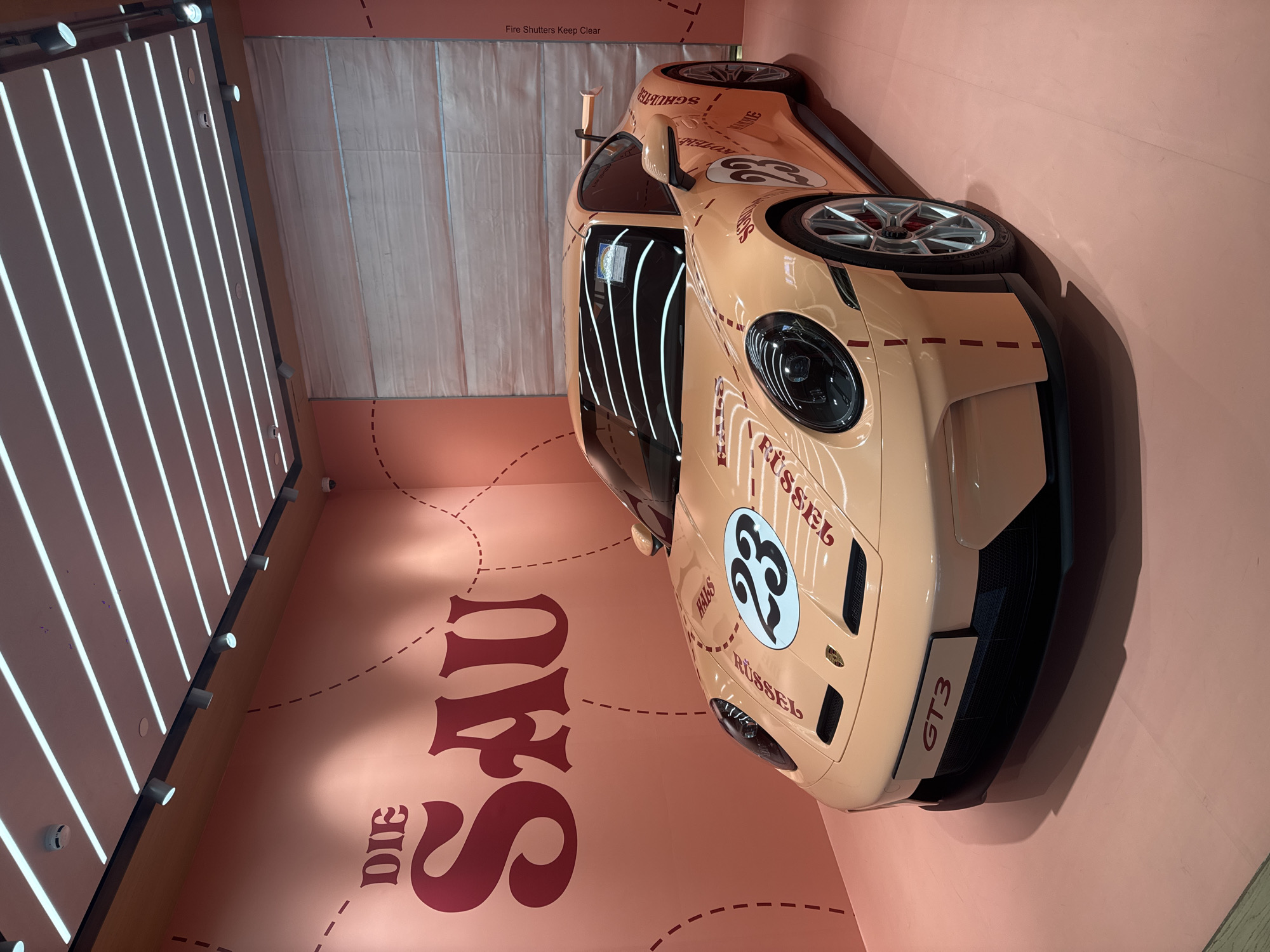 The image shows a Porsche GT3 car on display at an event in Singapore. The car is painted in a light orange color and has the number 23 on the hood. The words "DIE SAU" are written in large letters on the wall behind the car. The location is likely an exhibition or car show in Singapore. There is a wall with vertical lighting bars to the left of the vehicle. Another wall with "Fire Shutters Keep Clear" is behind the car.