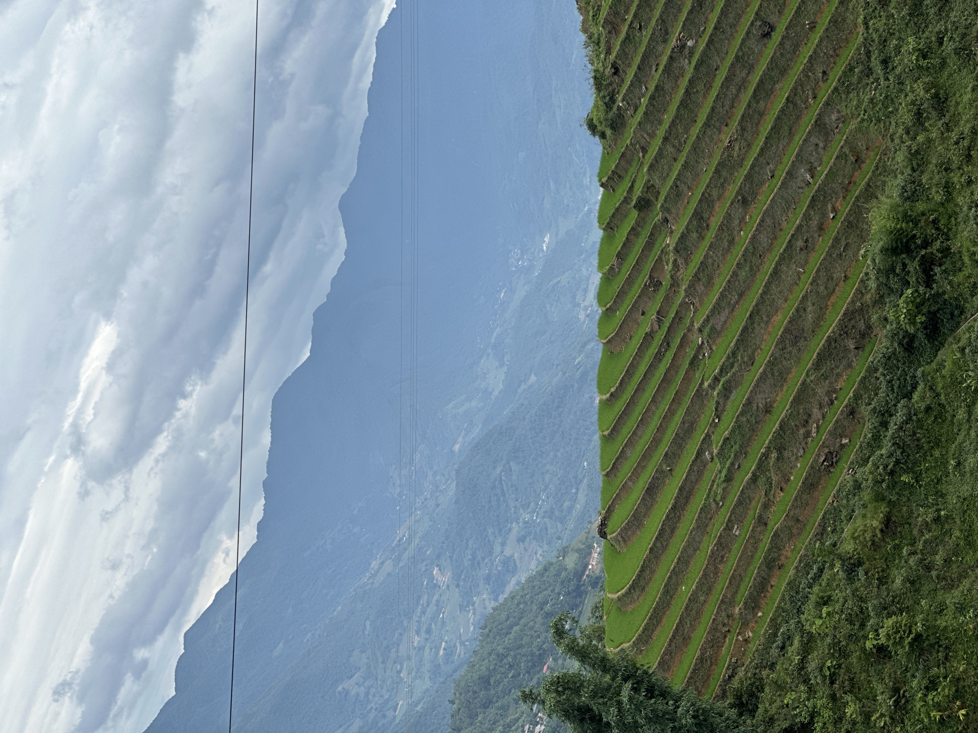 The image captures terraced rice fields on a hillside in Ban Ho, Vietnam, under a sky with partially cloudy conditions. The fields are layered, creating parallel green lines that contrast with the surrounding vegetation. The left side of the image reveals mountains obscured by haze and clouds. Electrical lines cut across the view towards the sky.