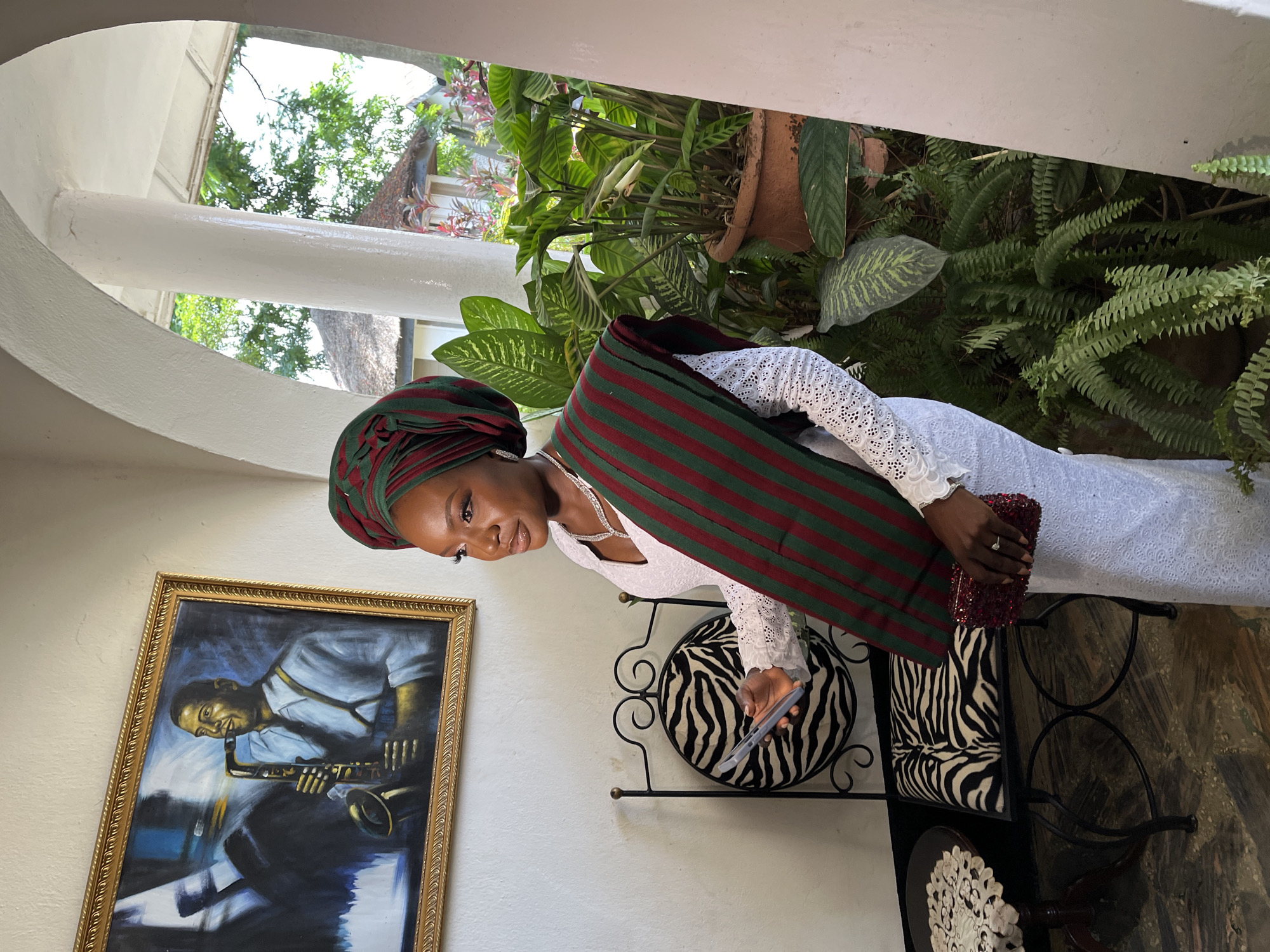 In the city of Bar Jirgi Summa, Nigeria, a woman stands in an indoor-outdoor space. She is wearing a white lace dress and a headwrap and sash with green and maroon stripes. A gold-framed painting of a man playing a saxophone hangs on the wall behind her. To her left is a black metal chair with a zebra-print cushion. In her right hand, she holds a smartphone, and in her left, a red sequined clutch. Lush green plants fill the space to her right, with some of the greenery visible through an archway leading to an outdoor area.