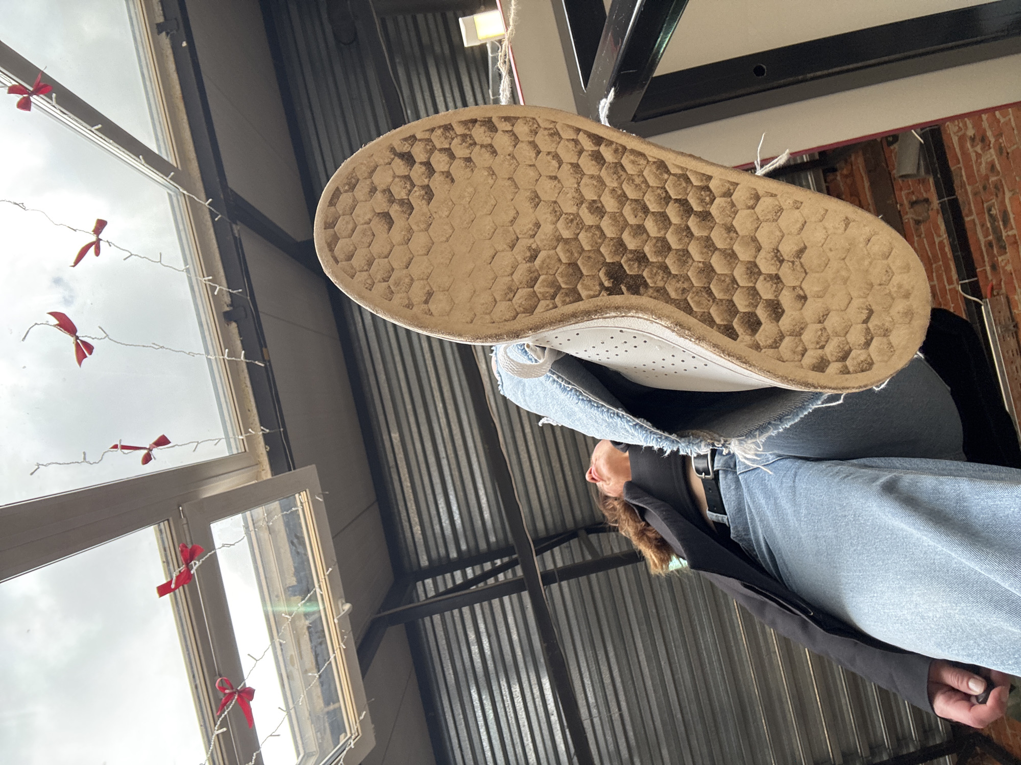 This image, taken in Chaikovsky, Russia, features a low-angle perspective looking upwards towards a person. The most prominent subject is the sole of a white sneaker, which is light brown, dirty, and features a distinct honeycomb pattern, occupying the upper central portion of the frame. The shoe's white upper, with perforated details, is also visible. Below the shoe, parts of a person are seen: light blue denim jeans with frayed edges, a dark belt, a dark long-sleeved top or jacket, and the side of their head with light brown hair, looking upwards.
The scene appears to be indoors, likely in a space with an industrial or rustic-chic aesthetic. To the left, a large window reveals an overcast, bright daytime sky, suggesting a cloudy day. White string lights decorated with small red bows are draped across the window, adding a decorative element. In the background, metal beams and corrugated metal siding suggest the building's structure, possibly a ceiling or wall. A reddish-brown brick wall is also visible towards the right, further defining the setting. No specific activity or event is clearly depicted beyond the static pose for the photograph. There is no visible text in the image.