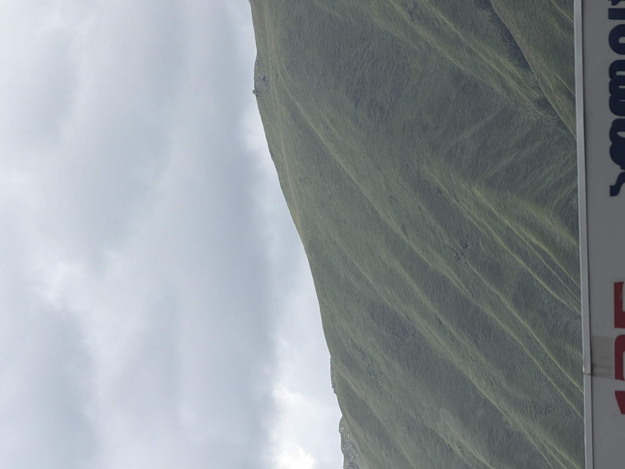 The image depicts a natural landscape featuring a vast, green, and rolling mountain range under a cloudy sky. The terrain is characterized by smooth, grass-covered slopes that exhibit distinct parallel ridges, suggesting natural formations or possibly terracing. The top of the mountain ridge is uneven, with some rocky outcrops visible. In the foreground, a portion of a white structure with red and dark lettering is present, which appears to be a sign or part of a vehicle, though the text is largely obscured or incomplete. There are no people or animals visible in the scene. The environment is predominantly natural, with the mountain occupying the majority of the frame. The location is identified as being in Almasiani, Georgia.