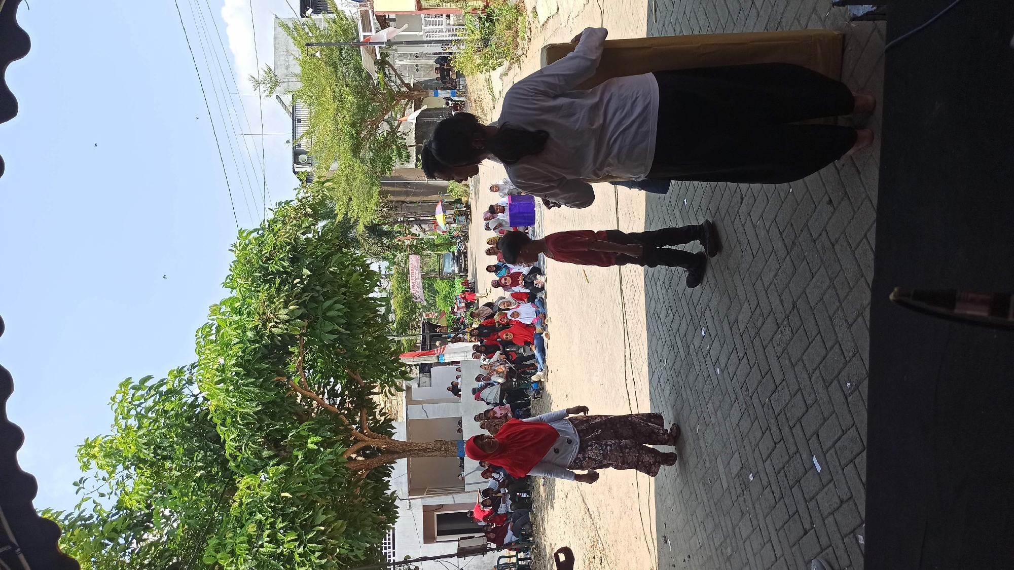 In the city of Galak, Indonesia, a group of people are gathered outdoors on a paved surface. In the foreground, a young boy in a red shirt and black pants stands on the right, facing left. To his left, a woman wearing a red hijab and a patterned skirt is walking. Behind them, a seated audience watches an event. The audience is composed of men, women, and children, many of whom are wearing red and white clothing, suggesting a festive or nationalistic occasion. Some individuals are seated on chairs, while others sit directly on the ground. The environment is sunny, with trees providing shade in the background. Buildings and a street with parked vehicles are visible in the distance, indicating an urban or semi-urban setting. A banner with text is also visible in the background. The overall scene depicts a community gathering or event.