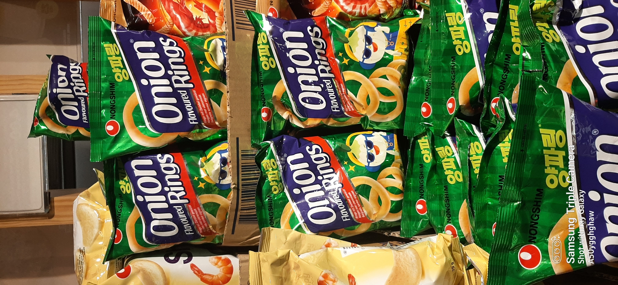 The image shows shelves stocked with bags of Nongshim Onion Rings, a popular Korean snack. The bags are predominantly green with blue and white lettering. Some bags are partially obscured by others, creating a layered effect. The lighting appears to be artificial, suggesting an indoor environment such as a supermarket or convenience store. The bottom of the image includes a Samsung Triple Camera watermark with the text "Shot with my galaxy A50yggghaw". The scene is devoid of people. There is no visual information in the image to suggest it is located in Dubai, United Arab Emirates.