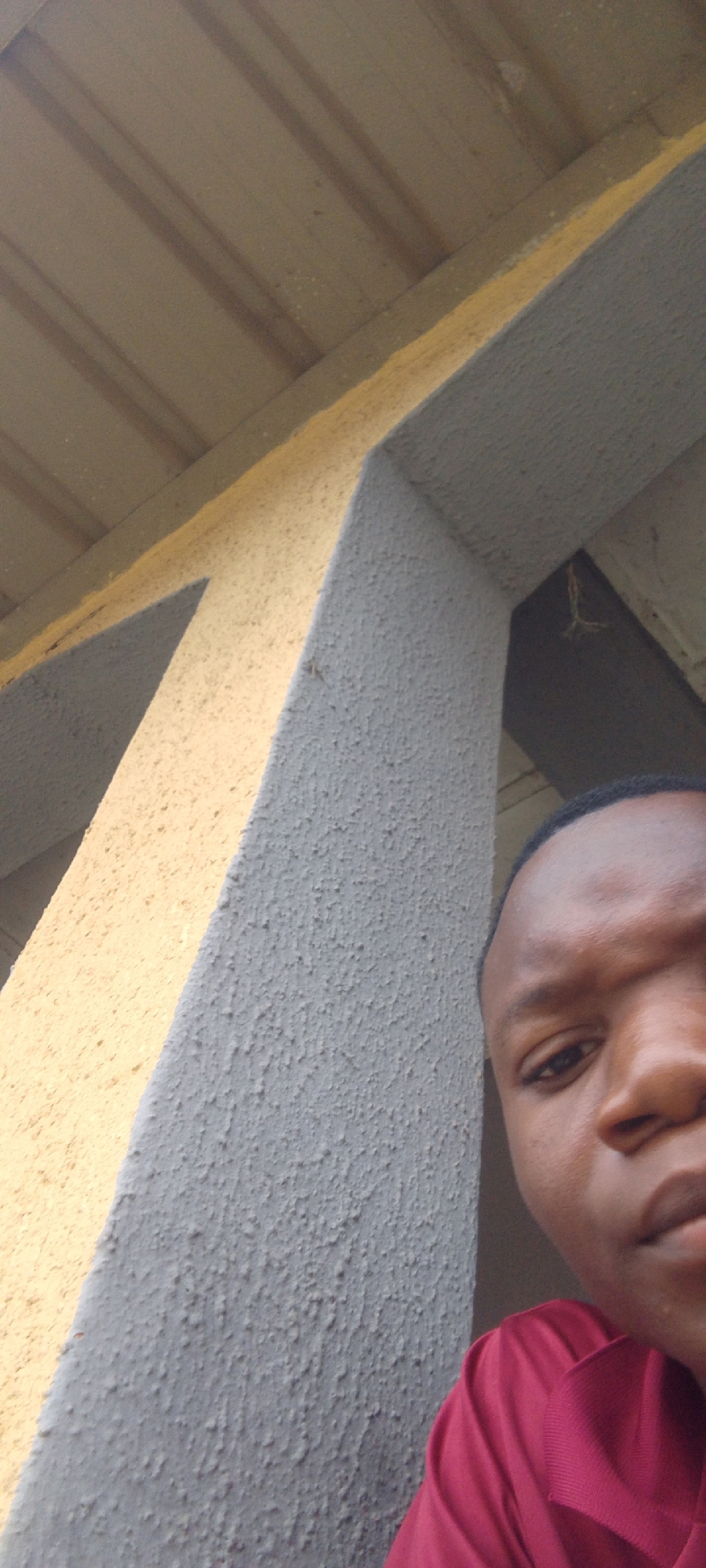 A close-up, low-angle shot shows the right side of a young Black person's face, mostly the eye, nose, and mouth, as they look towards the camera. They are wearing a dark red polo shirt. The person is positioned to the right of the frame, with a textured, grey concrete pillar to their left. Behind the pillar, a yellow, textured wall is visible, with a ribbed, light brown ceiling above. The scene appears to be outdoors, possibly under a covered structure. The lighting suggests it could be daytime. The overall mood is casual and direct, with the subject making eye contact. There is no visible text in the image. The provided location context is Suleja, Nigeria.