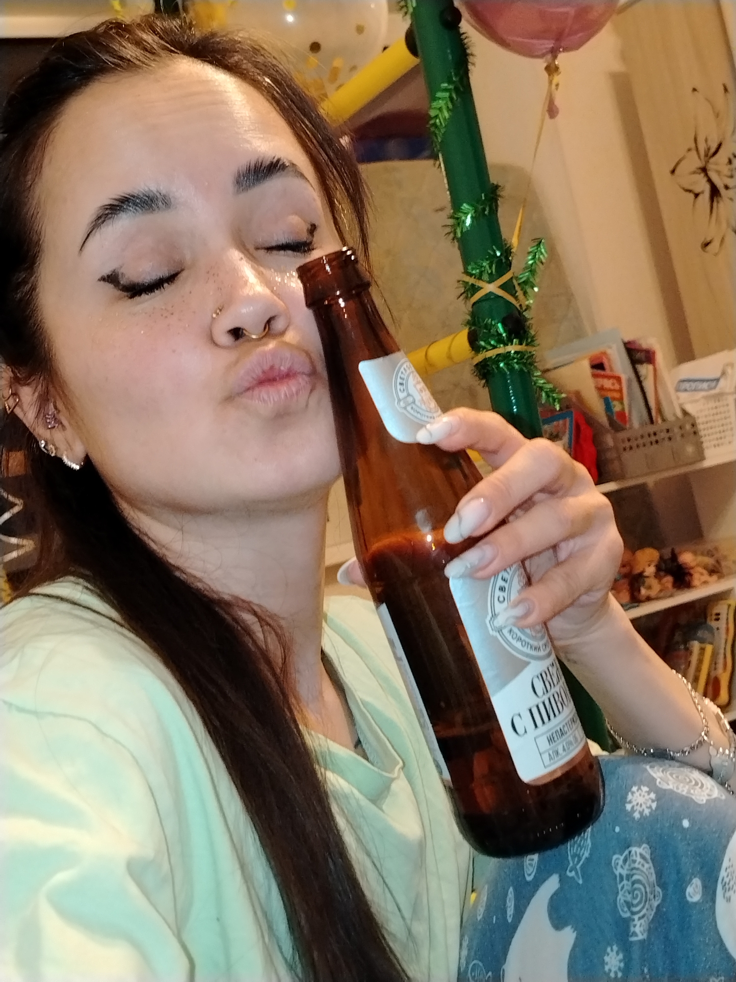A woman is taking a selfie with a bottle of beer. She has dark hair, fair skin with glitter, and is wearing light-colored pajamas. She has multiple earrings on her left ear and a nose ring. Her eyes are closed, and she is puckering her lips toward the beer bottle. The bottle is brown glass with a white label that reads "СВЕЖЕЕ С ПИВОМ" and "NEHACTEPOB". The label also indicates "ALK. 4.0% об." A pole wrapped in green tinsel is visible behind the woman, along with a pink balloon. A bookshelf with various items is in the background, and a flower print is on the wall. The beer is labeled as produced in Noviy, Russia.