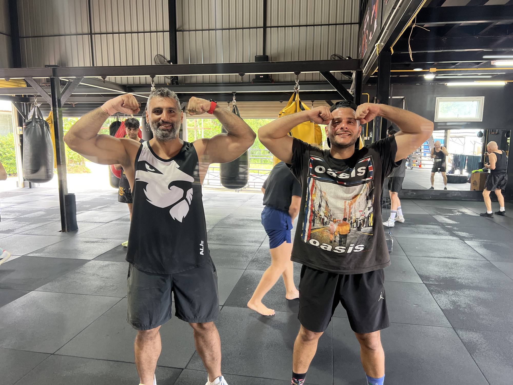 In a gym in Chalong, Thailand, two men are posing for a photo, flexing their biceps. The man on the left is wearing a black tank top with a white graphic of a stylized face. He has grey hair and a beard, and he is smiling. The man on the right is wearing a black t-shirt with a graphic of a street scene. He has short dark hair and is also smiling. Both men are wearing athletic shorts. In the background, there are punching bags, gym equipment, and other people working out. The floor is black rubber matting.