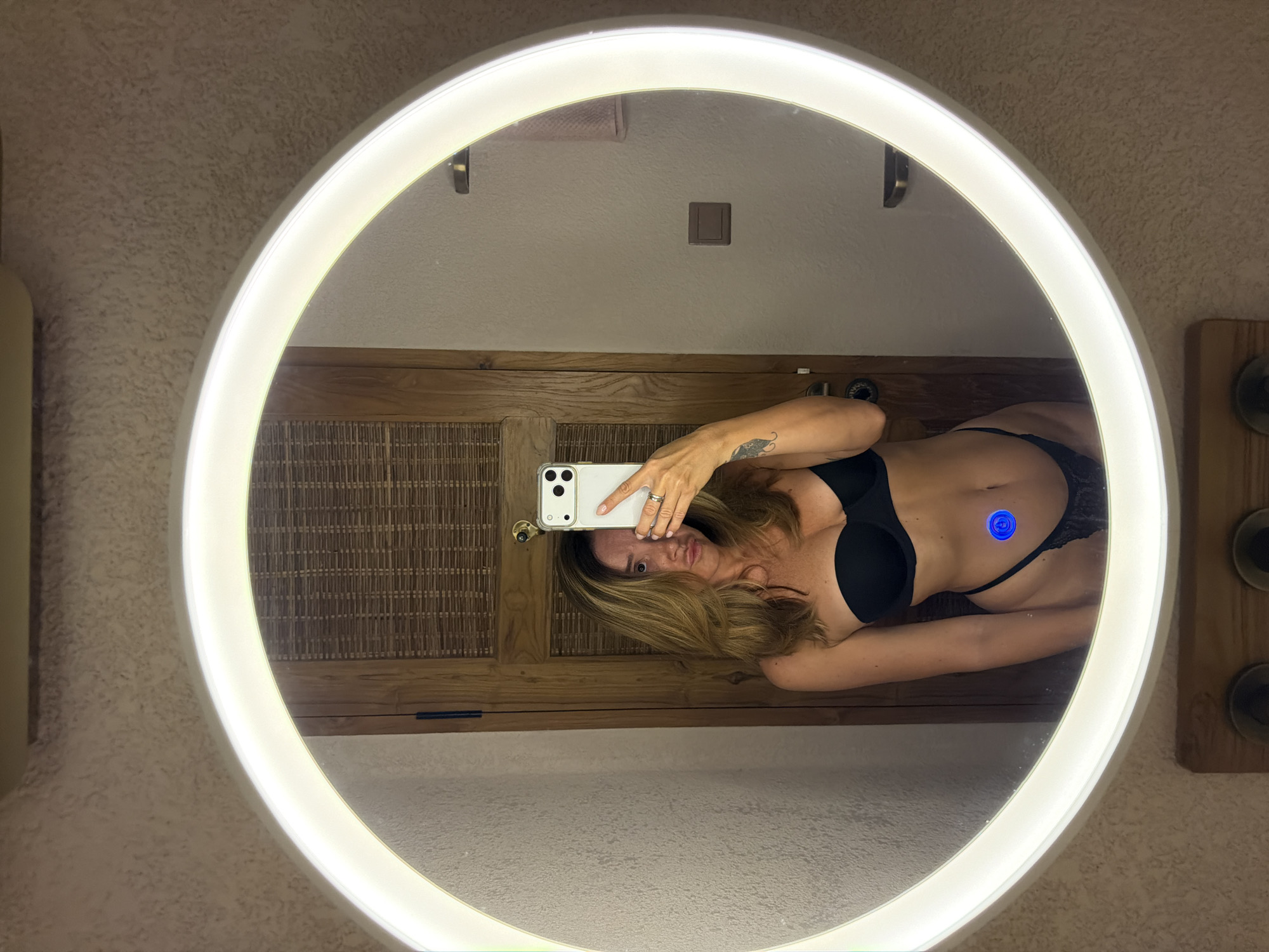 A woman is taking a mirror selfie in what appears to be a bathroom. She is wearing a black strapless bra and black underwear. Her hair is blonde and she has a tattoo on her left arm. In the reflection, a wooden door with a woven texture is visible behind her. There is also a circular mirror with a bright light around its edge. A blue circular sticker is on her stomach.