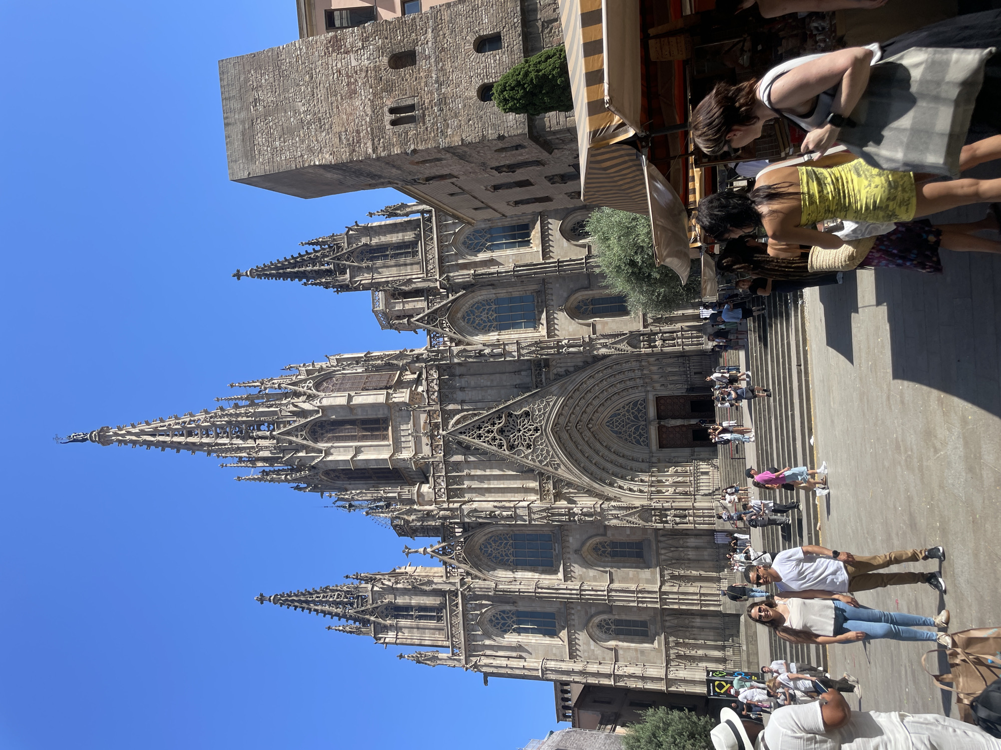 The image depicts a bright, sunny day in Barcelona, Spain, with the iconic Barcelona Cathedral as the prominent backdrop. Several people are visible in the foreground and on the steps leading up to the cathedral entrance. They appear to be tourists, some posing for photos, others browsing market stalls set up along the side of the street. The atmosphere seems relaxed and cheerful, indicative of a pleasant day out.
The architecture of the cathedral is intricate and grand, with soaring spires and detailed stonework, suggesting a historical and culturally rich location. The sunlight casts sharp shadows, indicating it's likely midday. The scene is bustling with activity, with people moving around and engaging in various activities. The market stalls suggest a commercial area where local goods or souvenirs might be sold. The clear blue sky further emphasizes the pleasant weather conditions. The overall impression is that of a vibrant tourist destination on a beautiful day.