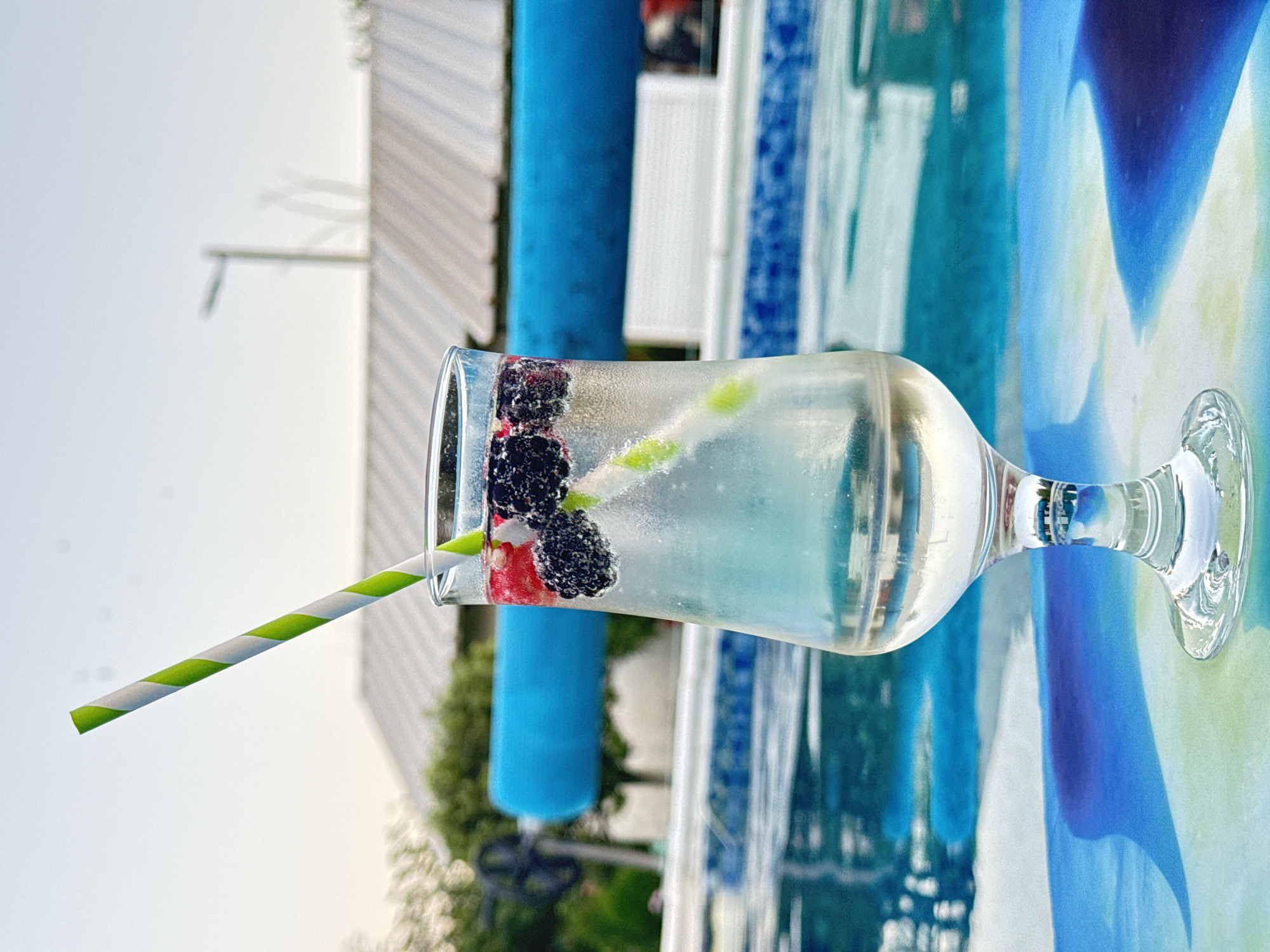 A clear glass of clear liquid with blackberries and raspberries and a green and white striped straw is placed on a table with a blue and green abstract pattern. In the background, a swimming pool with blue tiles is visible. A blue pool cover is stretched across the far end of the pool. Beyond the pool, there are trees and a building with a white roof. The sky is visible in the upper portion of the image. The scene appears to be outdoors, possibly in a backyard or a resort area. Bulgarovo, Bulgaria is not explicitly depicted in the image, and there is no information to confirm its location.