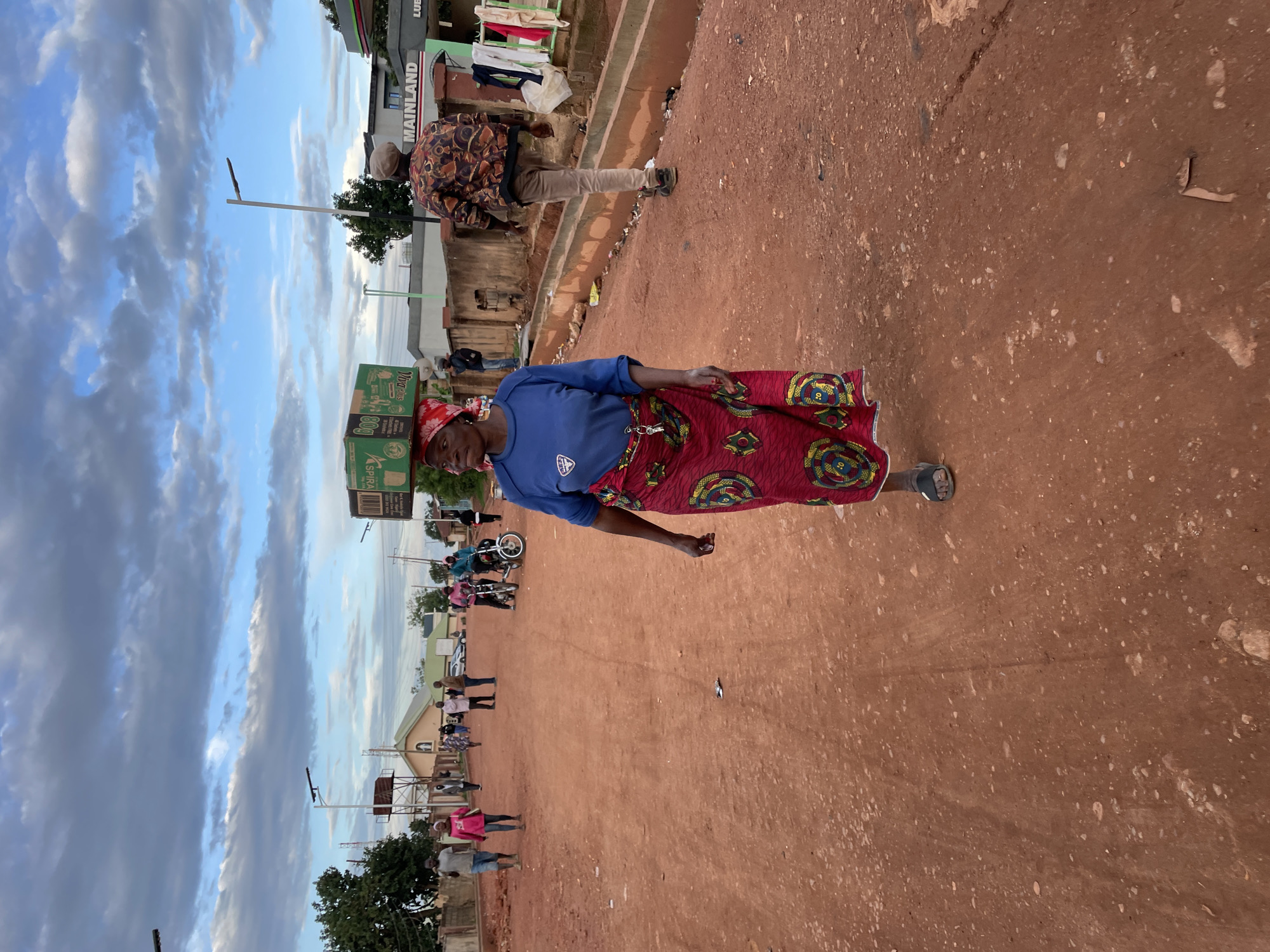This image captures a street scene in Gura Top, Nigeria, under a partly cloudy sky. The foreground is dominated by a mature woman, positioned slightly left of center, walking towards the viewer's left. She wears a blue long-sleeved shirt, a red and yellow patterned traditional skirt, and dark sandals, with a red headscarf. On her head, she carries a stack of green boxes, bearing the label "Viva Plus." A set of keys hangs from her left hip. The ground is an unpaved, reddish-brown dirt road, showing visible small stones and faint tracks.
In the mid-ground, multiple individuals are present. On the left side of the road, several people walk; one man is in a grey shirt and shorts, another person wears a pink top. Behind them, a building with a light-colored gabled roof and walls is visible, alongside trees and a streetlight. On the right side of the road, a man in a patterned jacket and light-colored pants walks away from the viewer. Behind him is a building displaying the word "MAINLAND," and clothes are hanging to dry on an adjacent structure. Further down the road, two individuals are seated on motorcycles. Another streetlight is visible on the right.