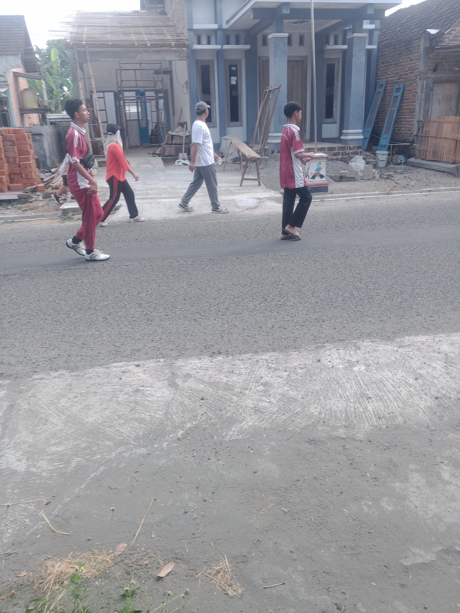 In Lembah, Indonesia, on a paved road with a concrete shoulder, four individuals are walking. The first person, a male wearing a maroon and white polo shirt and maroon pants, is in the foreground. To his left, a person in an orange shirt and black pants walks on the sidewalk. Further down the road, a male in a white t-shirt and grey pants walks, followed by a male in a maroon and white polo shirt and black pants. The background shows a building under construction, with exposed framing and stacked bricks, and residential structures.