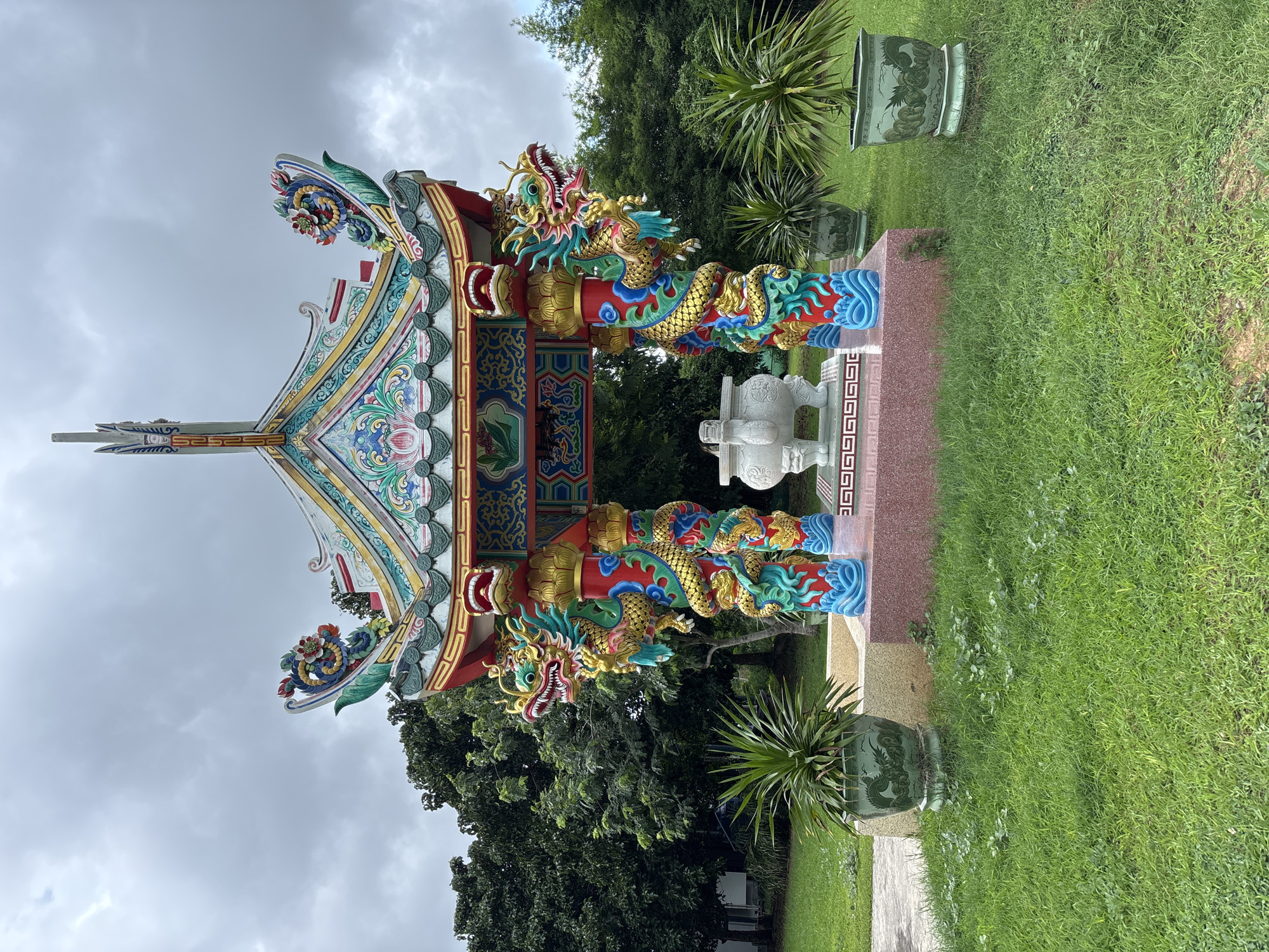 A vibrant, ornately decorated Chinese-style shrine stands in a grassy area under a cloudy sky. The shrine is supported by four colorful pillars, each featuring a detailed dragon sculpture. The roof is multi-tiered and adorned with intricate patterns and figurines. In front of the shrine, a large, traditional urn rests on a pedestal. Two potted plants with spiky leaves flank the shrine, placed in decorative green pots. The surrounding environment consists of lush green grass and dense trees in the background. The shrine appears to be located in the city of Chalong, Thailand.