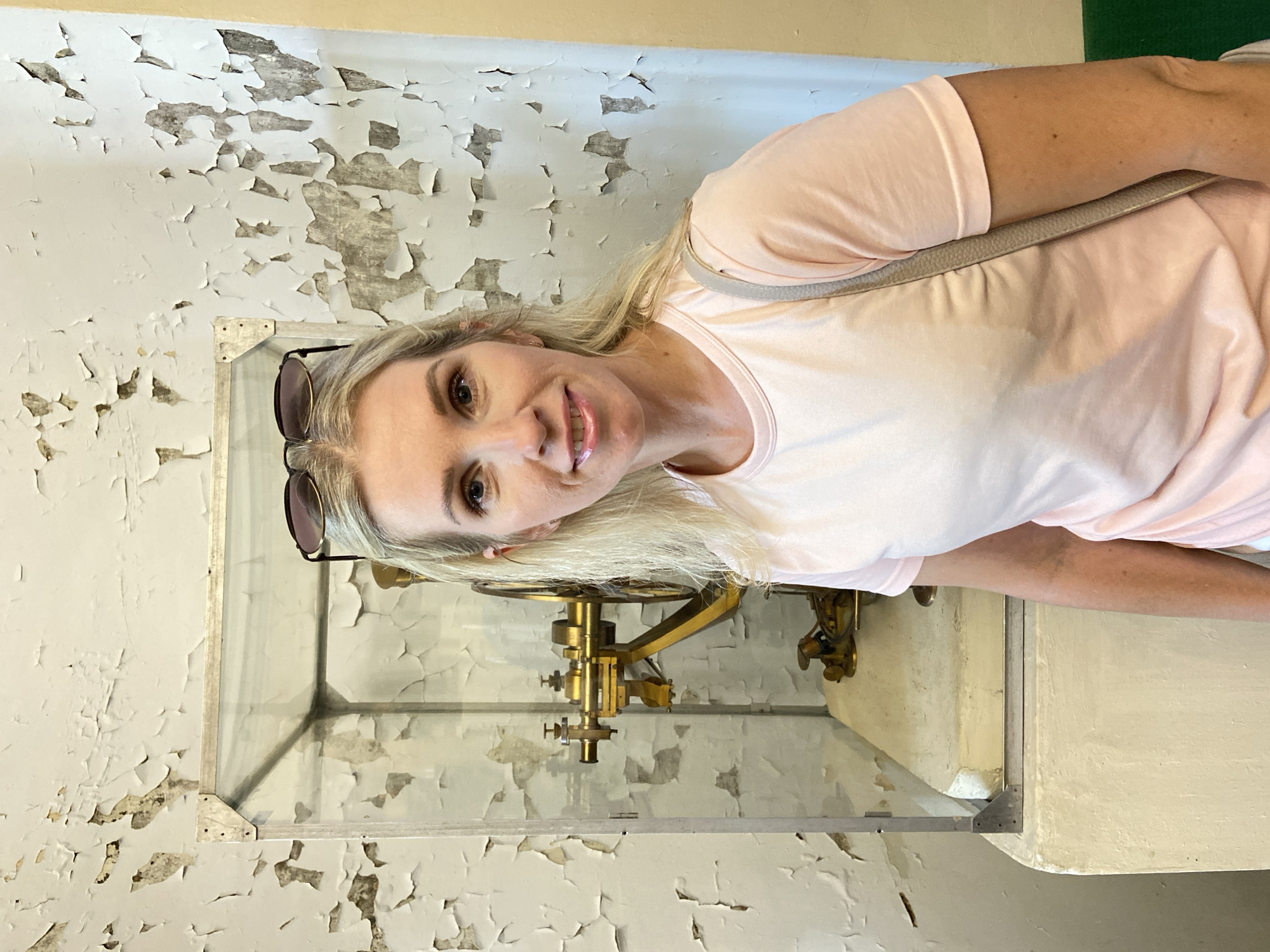 A blonde woman with sunglasses on her head stands in front of a display case containing a brass scientific instrument. The walls in the background are white with peeling paint, and the floor is a light yellow. The woman is wearing a pink t-shirt and has a grey purse slung over her shoulder. The scientific instrument appears to be a type of telescope or measuring device, with multiple adjustable parts and a circular wheel. The display case is made of glass with a metal frame. The overall impression is of a museum or historical exhibition setting.