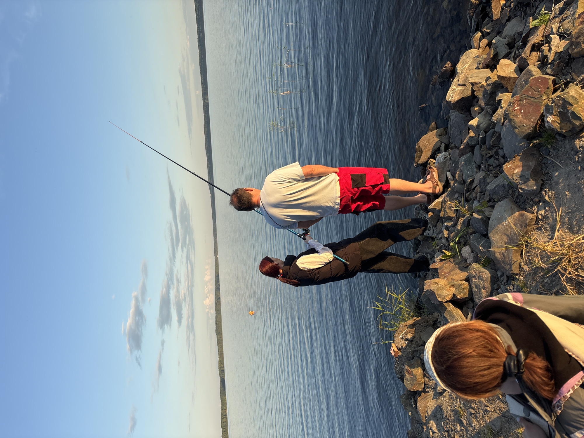 Three people are depicted on the rocky shore of a wide body of water, likely a lake, in Shuiskaya Chupa, Russia. The sky is clear with a few wispy clouds, indicating it is likely late afternoon or early evening. A man in red shorts and a white t-shirt stands facing the water, holding a fishing rod. Beside him, a woman wearing a black vest over a white shirt and black pants also holds a fishing rod, with her hand near the man's. In the foreground, another person, viewed from behind, with reddish-brown hair pulled back in a ponytail, is looking out towards the water. The water is calm and reflects the blue of the sky. A distant, treed shoreline is visible across the water. The foreground is covered in various sized rocks and some sparse vegetation.