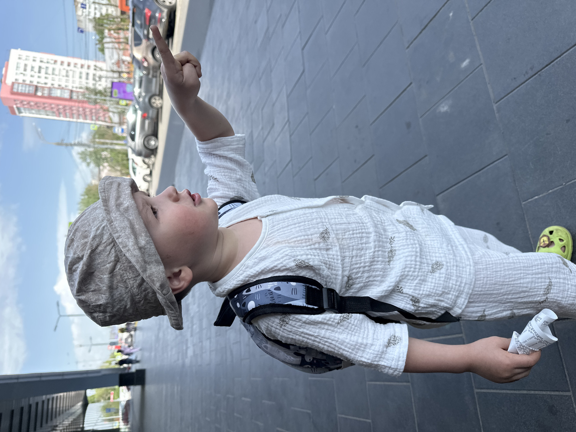 A young child in a light-colored patterned shirt and pants is standing on a sidewalk in Nizhny Novgorod, Russia. The child is wearing a cap and a backpack. The child is pointing upwards with their right hand and holding a crumpled piece of paper in their left hand. In the background, there are cars, a tall building, and streetlights. The sidewalk is made of gray rectangular tiles.