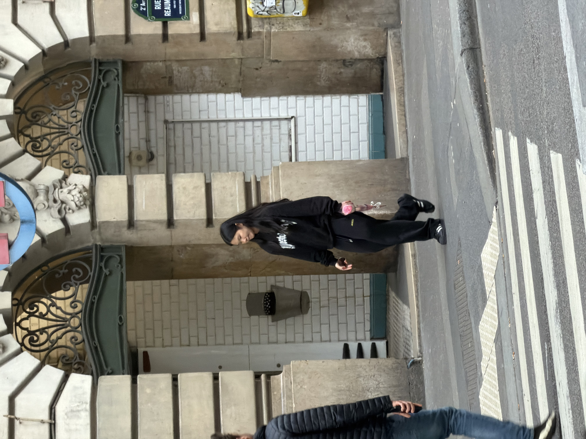 The image shows a street scene in Paris, France. The background includes a Metro entrance with the distinctive Art Nouveau wrought iron, and a street sign indicating "Rue Réaumur." A person wearing a black outfit is walking on the sidewalk, holding a pink flower. Another person wearing a jacket and jeans is partially visible at the bottom of the frame, also walking. A crosswalk is painted on the street. The sidewalk is concrete, and the wall of the Metro entrance is tiled.