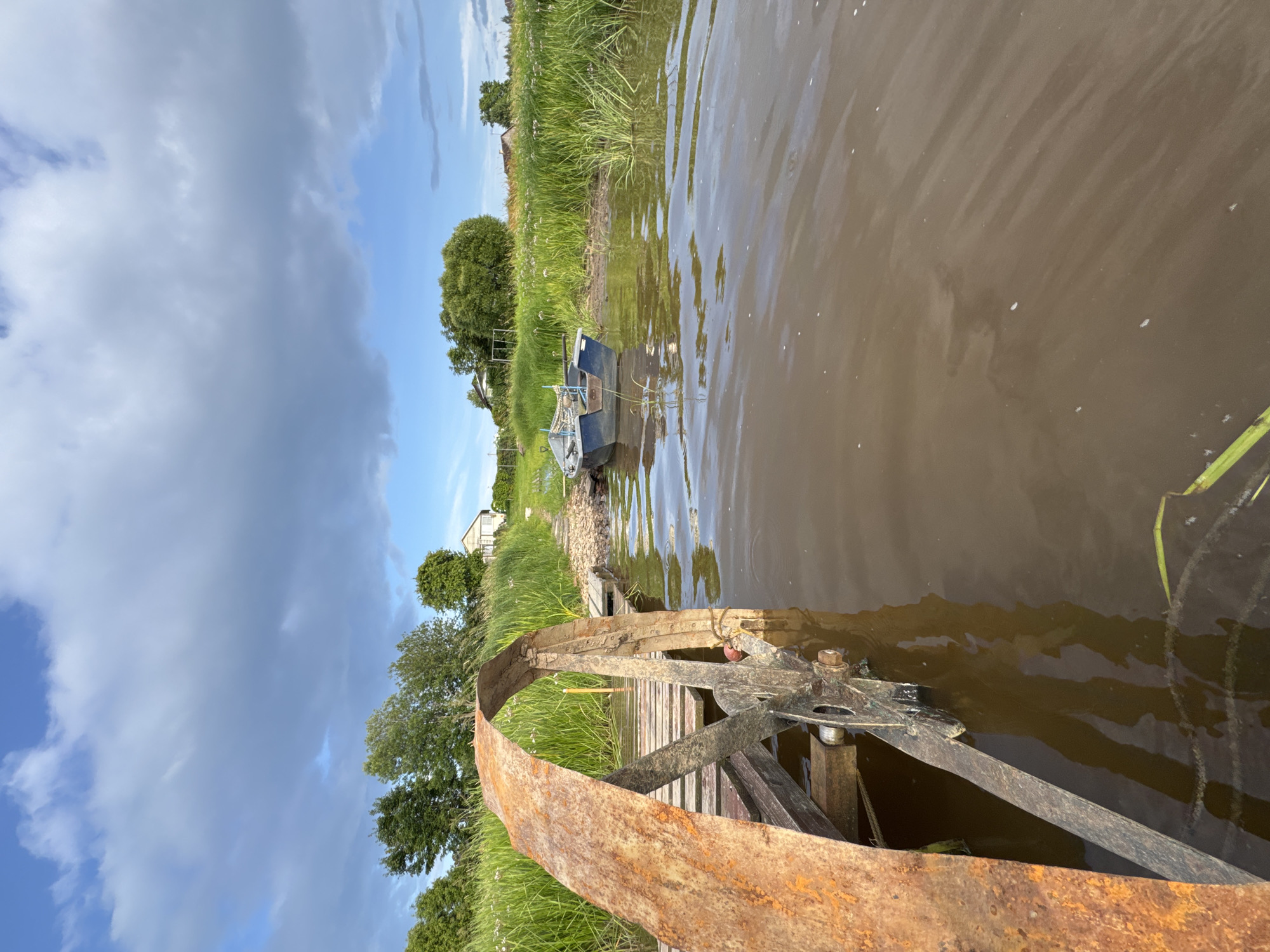 A low-angle, long shot captures a rural waterside scene with a rustic wooden pier and a rusty water wheel in the foreground. The pier extends from the left edge of the frame into muddy brown water. A large, curved, rust-colored metal wheel is attached to the pier and partially submerged. Beyond the pier, a small, dark blue boat rests on the muddy bank. The background features tall, green grass and reeds, a few scattered trees, and white houses with pitched roofs under a cloudy blue sky. The overall impression is one of a serene, undeveloped rural landscape.