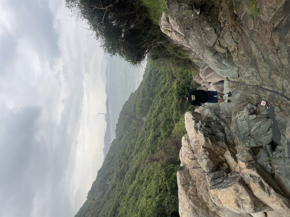The image captures a steep coastal incline in the city of Ninh Hòa, Vietnam, under a heavily overcast sky. In the immediate foreground, a single adult male, viewed from behind, stands on a complex formation of large, irregular rock boulders ranging in color from light tan to dark grey-green. He is dressed in a black long-sleeved shirt with a multi-colored logo on the back, blue shorts, dark socks, and dark shoes; a tattoo is visible on his right calf. He faces downwards, observing the terrain.
Below his position, the hillside descends steeply, covered with dense green vegetation, primarily shrubs and trees. A narrow, winding road or path is visible cut into the vegetated slope. To the right, the terrain meets a body of water, likely the ocean, which appears choppy. In the far distance across the water, an industrial complex or power plant is identifiable by several large structures, including at least one tall smokestack emitting a light-colored plume. A thick, black hose or cable extends diagonally down the rocky foreground from the lower right towards the center. A small, red and white object is situated on the rocks in a shallow pool of water in the lower-left foreground.