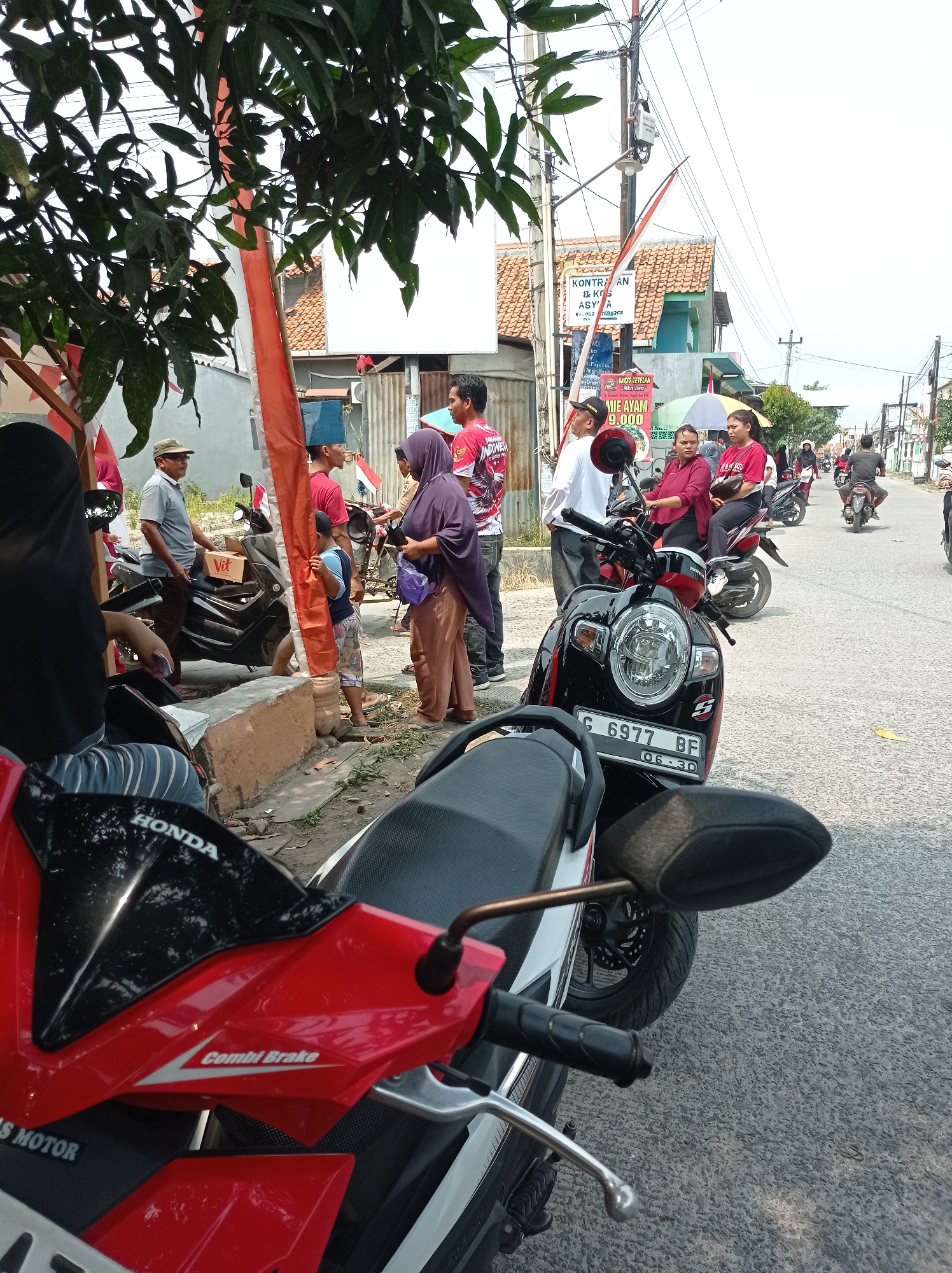 The image depicts a street scene in Kramat, Indonesia, on a sunny day. Several people are gathered near parked motorcycles. In the foreground, a red and black motorcycle is partially visible, with "HONDA" and "Combi Brake" branding on its body. Behind it, a black motorcycle with a license plate reading "G 6977 BF" is parked.
A group of individuals stands around the motorcycles. One man in a gray shirt and a cap is talking to someone out of frame. A woman in a purple hijab and brown pants is holding a purple bag. Another person, a child in a patterned shirt and shorts, is standing near a red and white Indonesian flag. Other people in red shirts are also present.
In the background, a street with more motorcycles and a few buildings is visible. A sign advertising "MIE AYAM 9,000" is prominent. Overhead, power lines crisscross the sky. The overall impression is a casual gathering on a street in a residential or commercial area of Kramat.