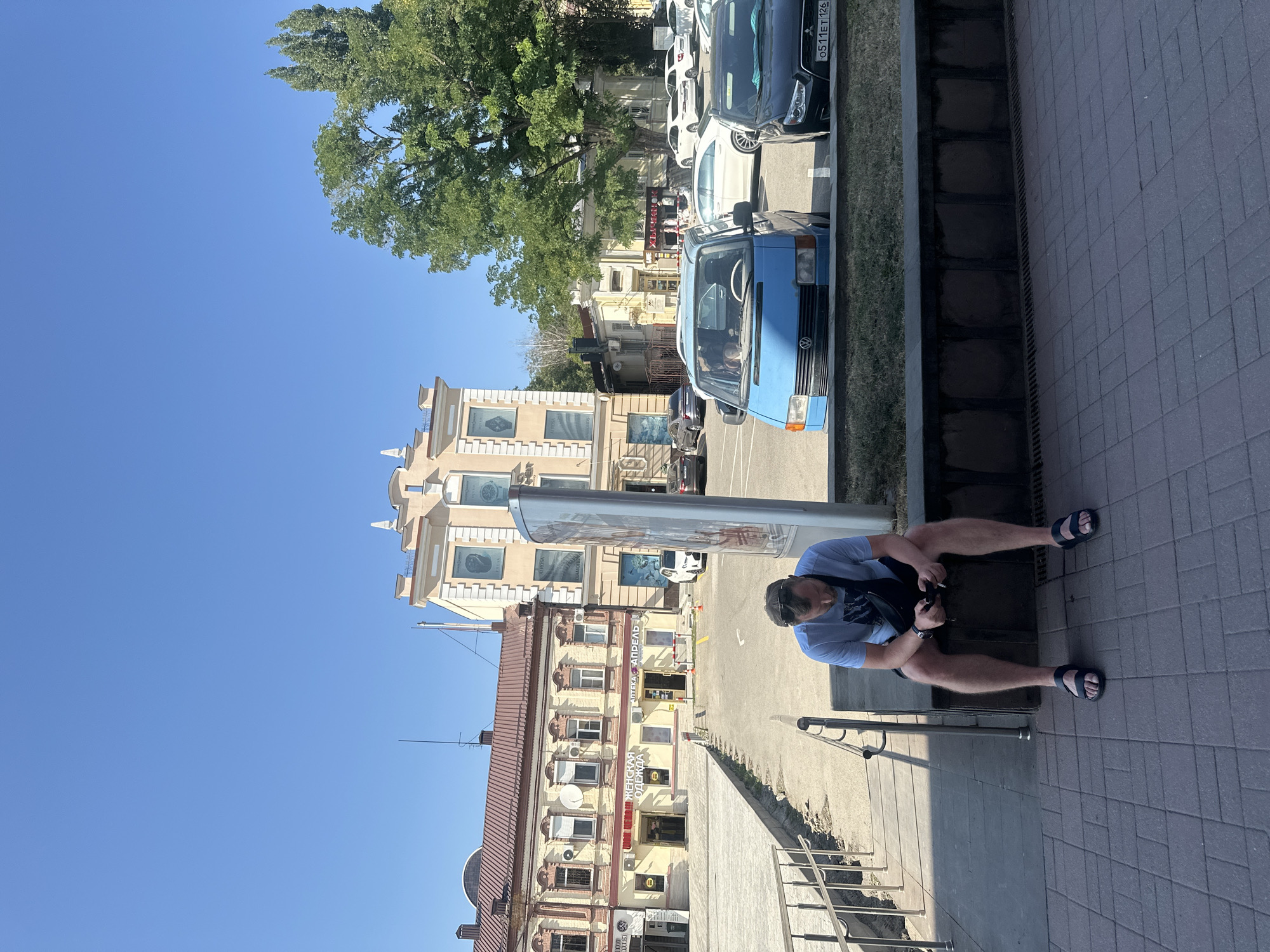 A man wearing a blue t-shirt, black shorts, and black sandals sits on a low wall outdoors in what appears to be a city square. He is looking down at his phone. A dark fanny pack is slung across his chest. He is positioned in the foreground, with buildings and parked cars in the background. The buildings are multi-story with a light-colored facade. A blue van and a black car are parked in front of the buildings. The sky is clear and blue, suggesting a sunny day.